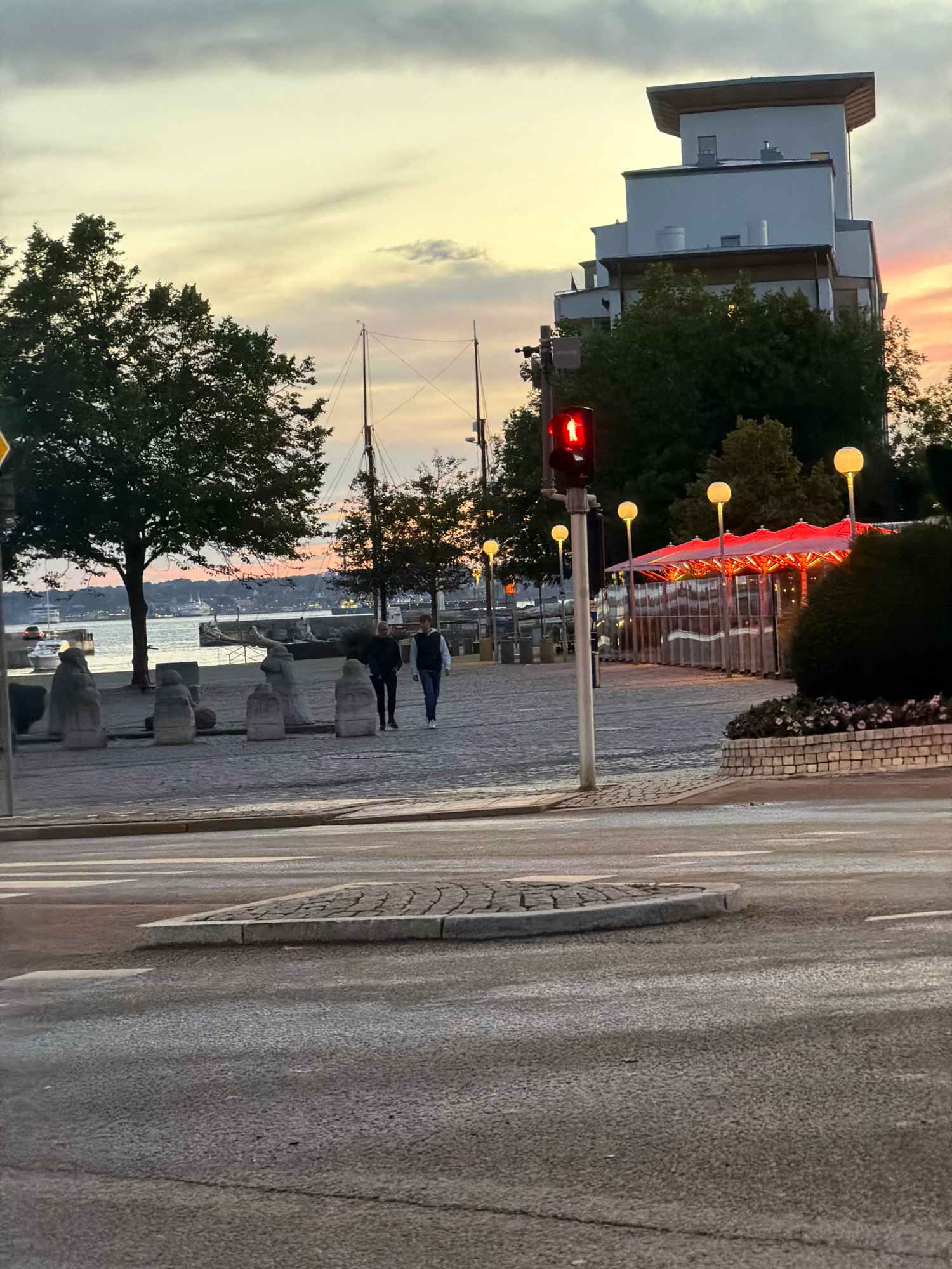 The image shows a street scene in Helsingborg, Sweden, at dusk. A traffic light displays a red signal for vehicles. Two men walk on a cobblestone path alongside a waterfront. In the background, a modern, multi-story building stands prominently, with trees and a red-canopied outdoor dining area in front of it. The sky displays soft, pastel colors indicative of sunset. The scene appears to be a pedestrian walkway or promenade next to a street and a body of water.
