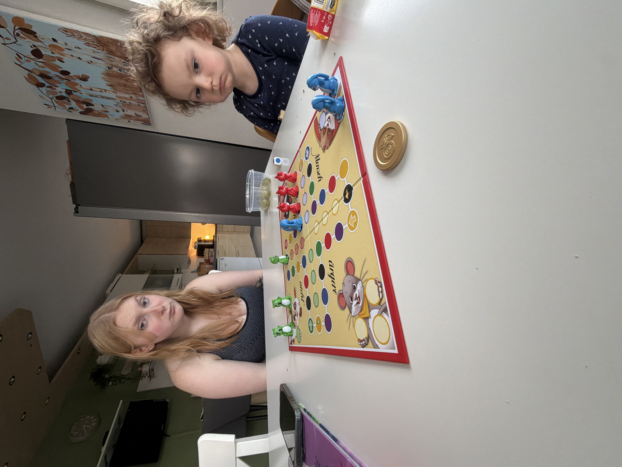 Two individuals are present in an indoor setting. A young child with curly hair is positioned at the top of the image near a table, seemingly participating in a board game. At the opposite side of the table, another person with long, light hair is also involved in the game. The board game is set on a white tabletop, with colored game pieces (red, blue, green), a coin, and a die visible. The game board features a yellow surface with a circular track, along with imagery of a mouse and the words "Mensch argere dich nicht". In the background, a kitchen area can be observed with a refrigerator and other appliances. The location is Trassem, Germany.