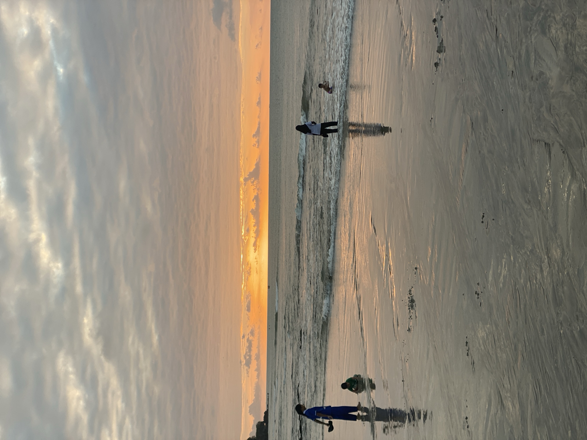 In Kuta Selatan, Indonesia, at sunset, four people are visible near the shoreline. On the left, a person in a blue outfit stands in the shallow water, holding a dark object. Nearby, a child in a green shirt is sitting on the wet sand, appearing to be playing. Further out in the water, another person in dark clothing stands, while a child in a swimsuit is swimming. The sky is filled with a gradient of orange and gray clouds, with the bright setting sun visible on the horizon. The wet sand reflects the light of the sky and the figures, creating a glistening effect. Small clusters of dark debris are scattered on the sand. The waves are gently rolling onto the beach.