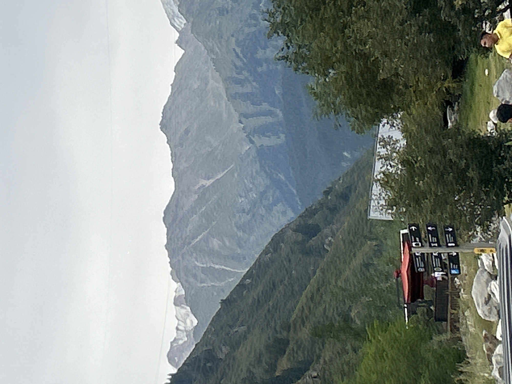 The image depicts a panoramic view of a mountainous landscape in the city of Almaty, Kazakhstan. The foreground features a cluster of trees and a red-roofed structure that appears to be an outdoor kiosk or information booth, with several directional signs. In the mid-ground, lush green slopes rise sharply, partially obscured by trees. The background is dominated by majestic, hazy mountains, with snow-capped peaks visible in the distance under a cloudy sky. Two individuals are partially visible in the lower right corner; one is wearing a yellow shirt and looking to the right, while another person's head and shoulder are visible, partially obscured by a rock. The overall scene suggests a scenic natural location accessible to visitors, possibly a park or a hiking trail entrance.