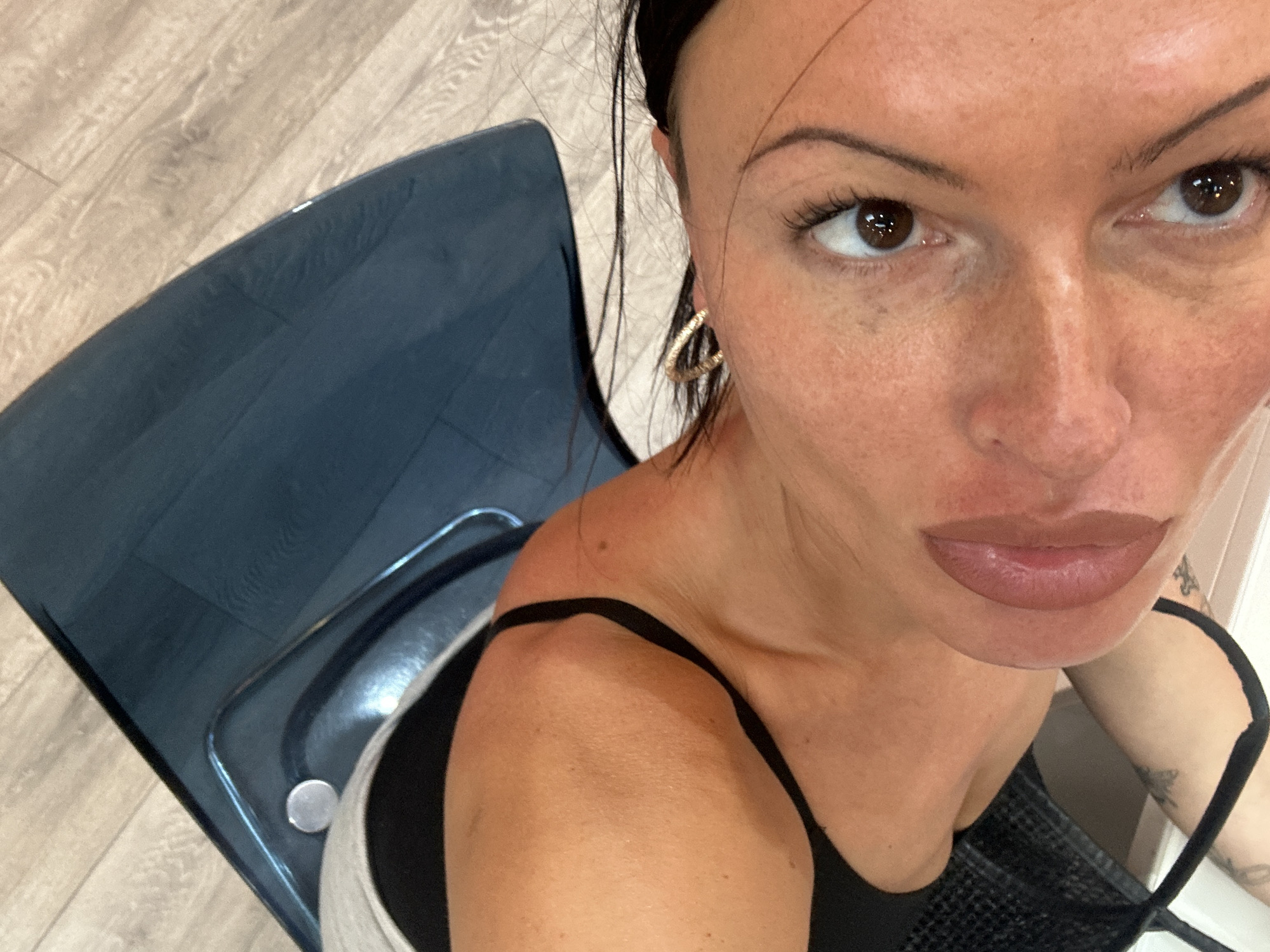 A high-angle, close-up selfie of a woman with dark hair pulled back from her face. She has light brown eyes and fuller lips. She is wearing gold hoop earrings and a black tank top with thin straps. Part of a blue, transparent chair is visible to her left. The background shows light-colored wooden flooring. A tattoo is slightly visible on her upper arm. There is no indication of this image being taken in Tunbridge Wells, United Kingdom.