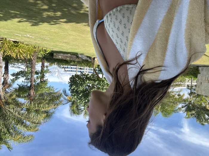 The image shows a female individual, visible from the mid-torso up, in profile, facing right. She has long, dark brown hair and wears a light-colored bikini top with a subtle pattern. A yellow and white striped towel is draped over her shoulders. The setting is outdoors in Marina Maroma, Mexico, under a clear blue sky with scattered white clouds. The foreground and midground consist of a well-maintained green lawn with shadows present. Several palm trees with green fronds and other tropical vegetation are visible on both sides of the frame and in the background. Modern, multi-story buildings with light-colored facades, consistent with a resort complex, are visible in the distant background. No other individuals or direct interactions are depicted.