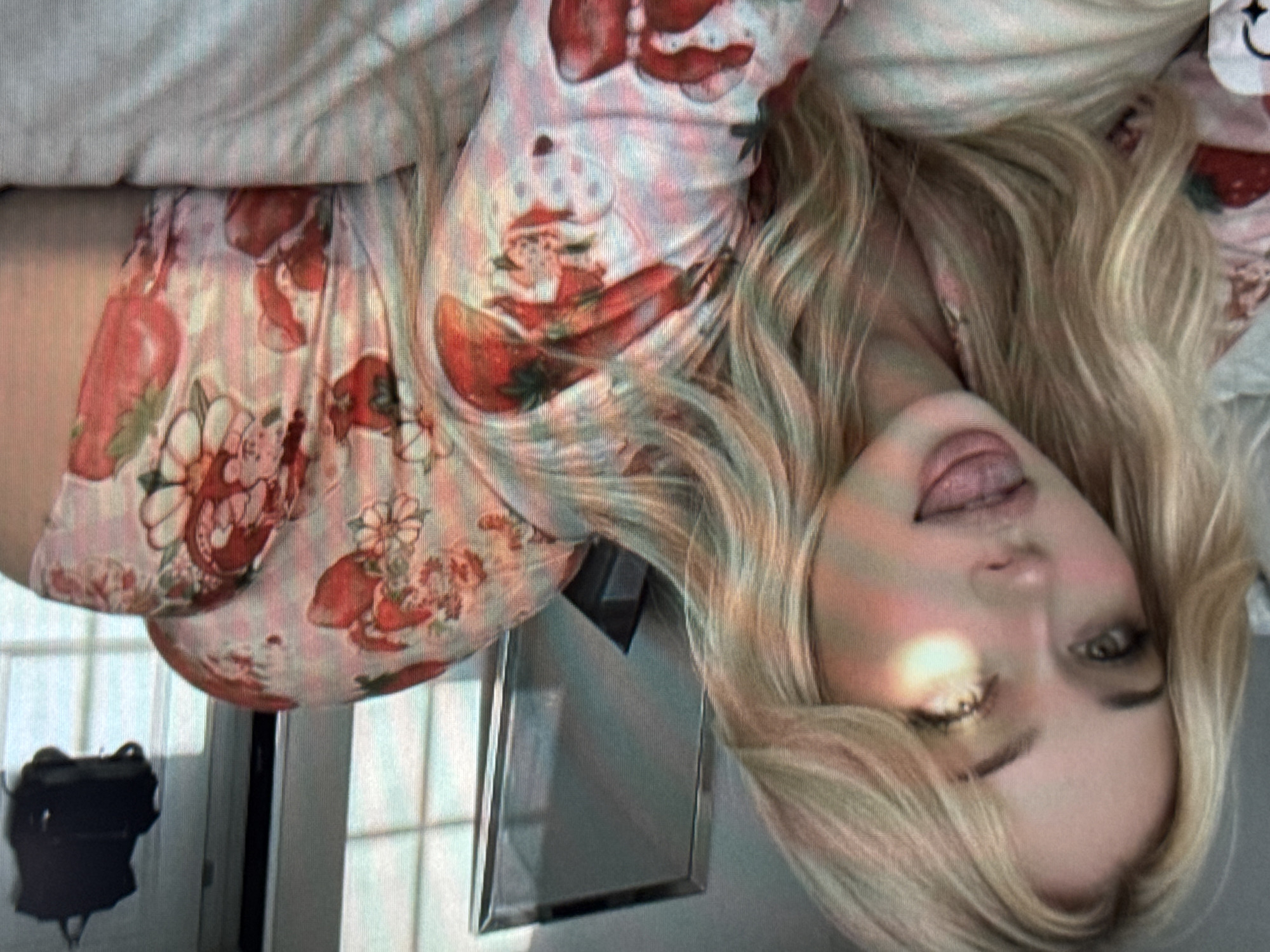 The image displays a young woman with long, wavy blonde hair, looking directly at the camera. She is lying down, with her head slightly tilted and her tongue subtly out, conveying a playful or relaxed expression. Her eyes are open, and there's a noticeable bright reflection or glare on her left cheek and hair.
She is wearing light-colored pajamas or loungewear featuring a distinctive pattern of red strawberries and small figures resembling Santa Claus in a sled. The fabric appears soft and comfortable.
The setting is indoors, likely a bedroom or living space. In the background, to the left side of the frame (relative to her body), a reflective surface, possibly a mirror or a framed item, is visible. Below this, a dark, irregularly shaped object, possibly a bag or piece of furniture, is partially seen. The overall lighting is soft and ambient. There is no visible text in the image.
