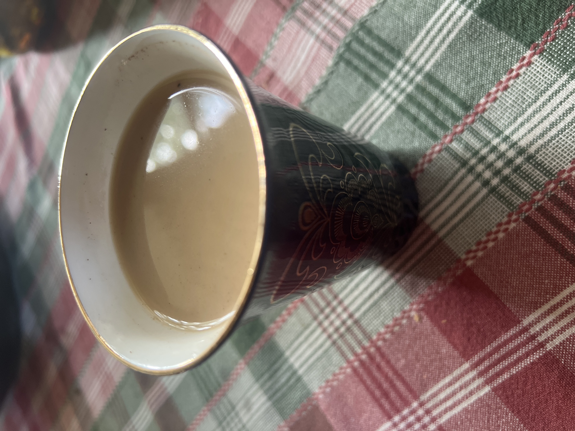A close-up, eye-level shot shows a dark, ornate cup filled with a light brown liquid, likely coffee or tea, with a golden rim. The cup sits on a red and green plaid tablecloth. The background is slightly blurred, but the overall lighting suggests an indoor setting. The image does not provide specific details to indicate a location in Norviliškės, Lithuania.