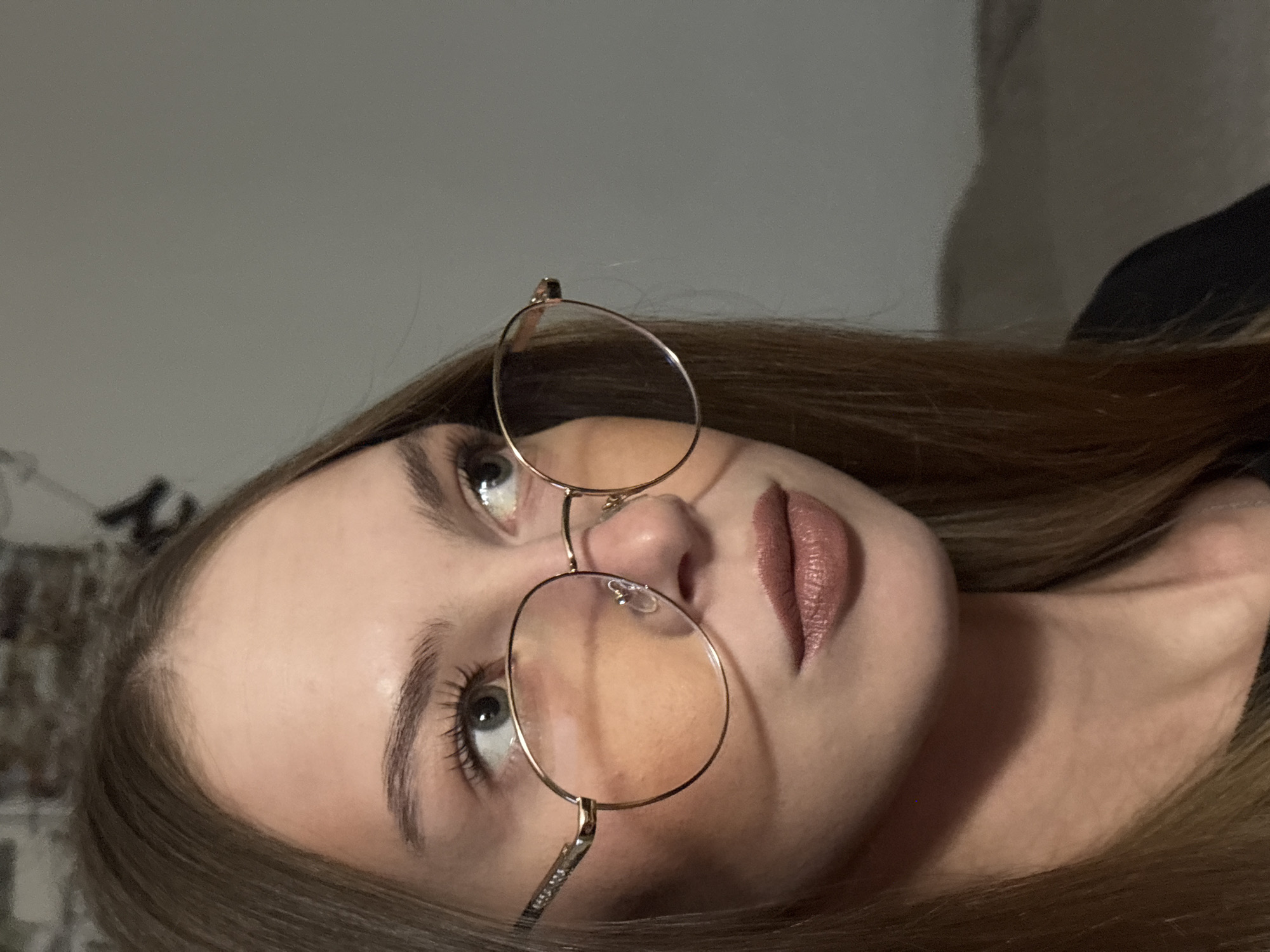 A young woman with long, straight brown hair, wearing round, gold-rimmed glasses with orange lenses, is shown in a close-up profile view against a plain gray background. She is looking to her right. Her makeup includes mascara and brown lipstick. The image provides no information to indicate a location in the city of Perm, Russia.