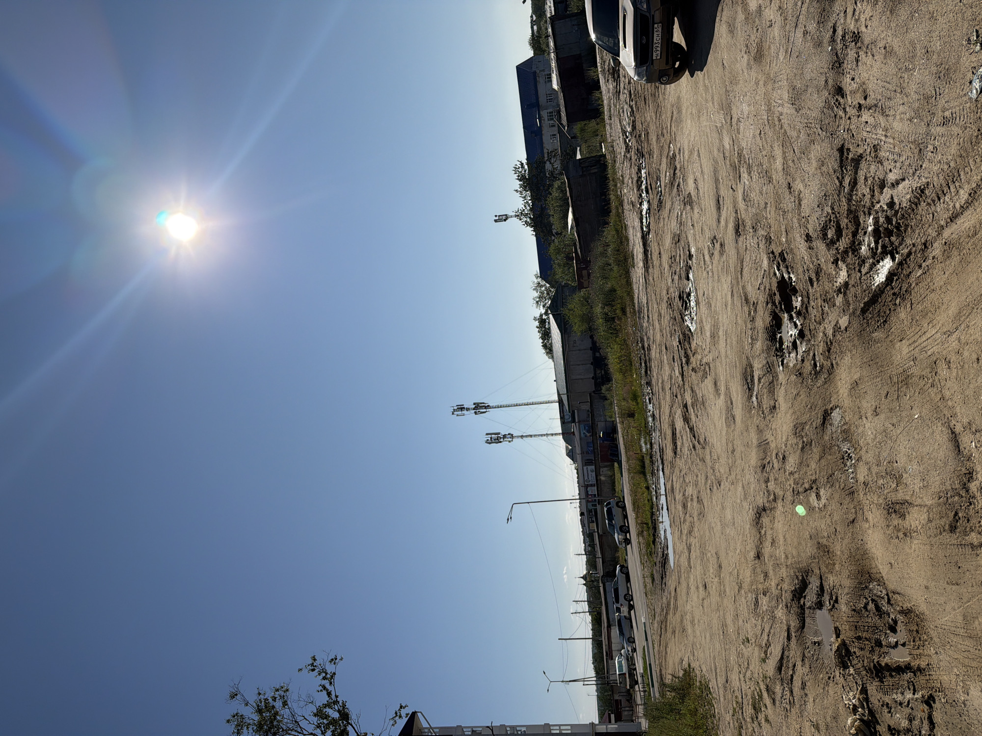 The image depicts a street scene in Okha, Russia, under a clear blue sky with the sun prominently visible and casting lens flares. The foreground is dominated by a muddy, rutted dirt road with several puddles. Tire tracks are visible in the mud. In the mid-ground, to the right, a light-colored SUV with a license plate beginning with "H993CH" is parked on the dirt. Further back, a paved road runs horizontally, with several cars parked alongside it. On the far side of the paved road, there are various buildings, including what appear to be industrial structures and smaller sheds. Two tall communication towers with antennas are prominent in the middle distance. To the left of the frame, a portion of a building with a white facade is visible. A bare tree branch extends from the upper left corner. The overall impression is one of an undeveloped or industrial area on a sunny day.