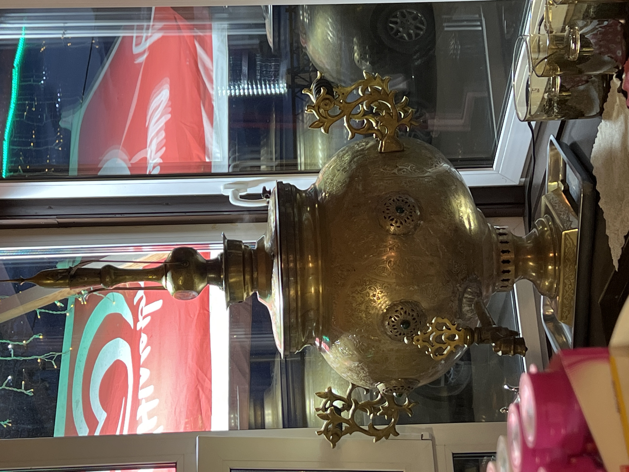 The image shows a vintage brass samovar positioned in front of a window. The samovar is ornate with intricate carvings and decorative handles. Behind the samovar, the window displays a red banner with white lettering and a green swirling design. The window also reflects some lights and what appears to be a car. In the foreground, to the left, several bright pink cylindrical objects are visible, possibly cups or containers. To the right of the samovar, on the windowsill, are several clear glass tumblers, some containing a reddish liquid or substance. The scene appears to be indoors, with a hint of outdoor elements visible through the window. The overall impression is of a traditional or cultural setting, potentially a cafe or shop in Listvyanka, Russia, known for its historical and cultural significance.