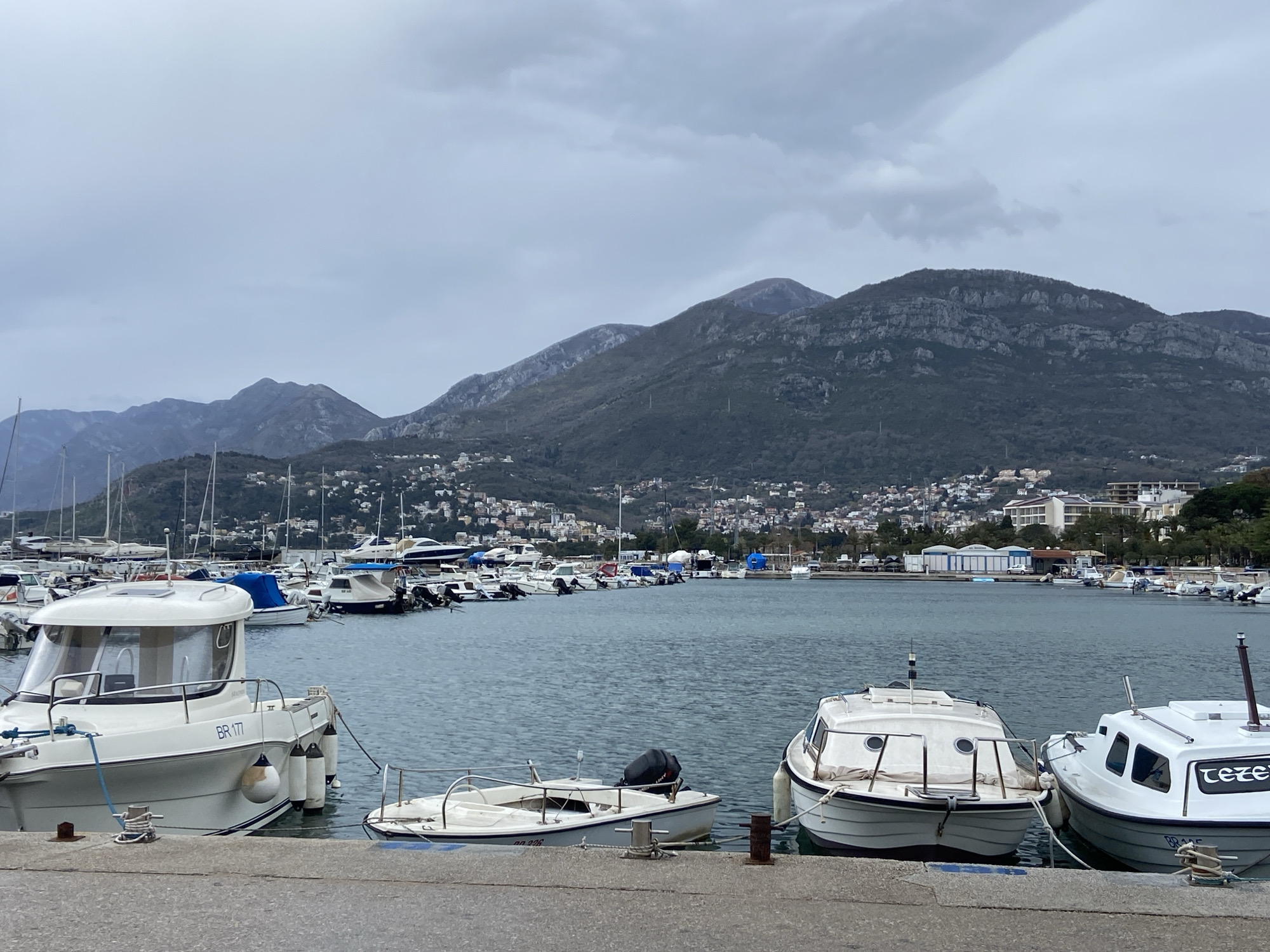 The image shows a marina in Bar, Montenegro on an overcast day. In the foreground are several white boats docked along a concrete pier. The boat on the left has "BR 171" printed on its side. To the right, a smaller boat with an outboard motor is also docked. Further along, two more white boats are moored side by side. The water in the marina is calm. Numerous other boats and yachts are docked in the background, stretching into the distance. Behind the marina, a town is nestled at the foot of a large mountain range under a gray sky.