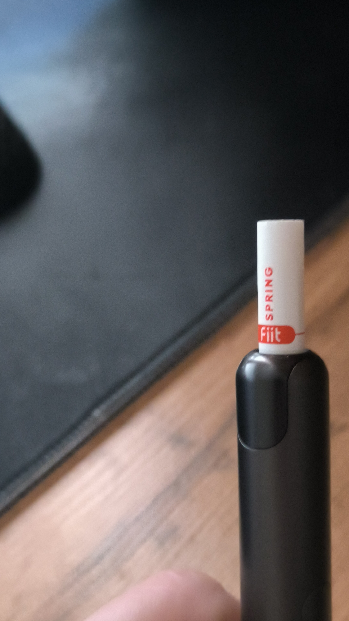 The image shows a close-up of a white and red "Fiit" HEET stick, possibly for an electronic smoking device, positioned on a wooden surface. The "Fiit" branding is visible in red text on a white background, with the word "SPRING" also printed vertically in red above it. A red line accentuates the branding. The stick is inserted into a black device, which takes up the lower portion of the frame. The background is blurred, showing a dark, textured surface on the left and a lighter, out-of-focus wooden surface on the right. A human finger is partially visible in the lower left corner, suggesting someone is holding or interacting with the device. The lighting appears to be artificial, possibly from an overhead source, creating soft shadows. There are no clear indicators of the time of day, weather, or specific emotions. The image is static and likely taken indoors, with the focus solely on the product and the device. Given the context, the object is likely being prepared for use or has just been used.