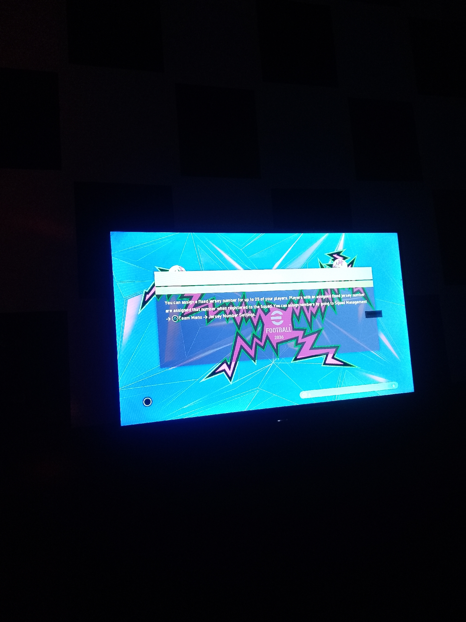 The image is a screenshot from a video game displayed on a screen. The game appears to be a soccer (football) game, as indicated by the text "FOOTBALL 2026" and the visual elements. The screen displays a bright blue background with a stylized, jagged pink and green graphic design. A white text box with information is overlayed on the screen. The text reads: "You can assign a fixed jersey number for up to 25 of your players. Players with an assigned fixed jersey number are assigned that number when registered to the Squad. You can assign numbers by going to Squad Management -> Team Menu -> Jersey Number Settings." The game's logo, a stylized "1" inside a circle, is visible. There is also a thin, horizontal progress bar at the bottom of the screen. The image does not contain any information related to the city of Serti, Nigeria.