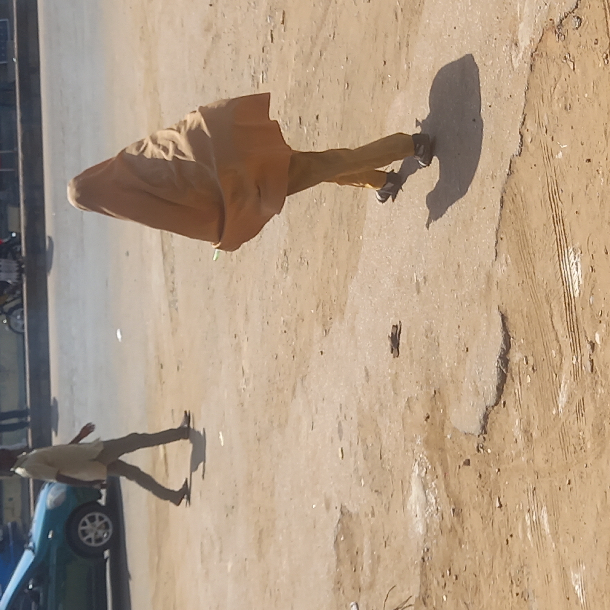 A person wearing a long, brown robe is walking on a dirt road in Sardauna, Nigeria. Another person, wearing light colored clothing, is walking on the road closer to the edge, beside a blue car. There are tire tracks and other debris on the road. The lighting is bright, casting shadows of the people and objects.