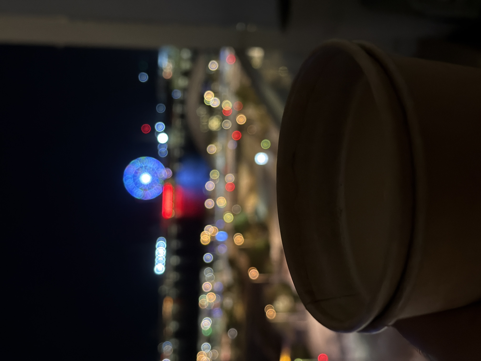A night shot from a low angle shows a disposable coffee cup in the foreground, held by a person's hand. The cup is light brown and slightly blurred. In the background, the city skyline is visible at night with bokeh lights. A large, illuminated Ferris wheel with blue lights is prominent in the center, with red and white lights scattered across the city. The scene appears to be from an urban waterfront area.