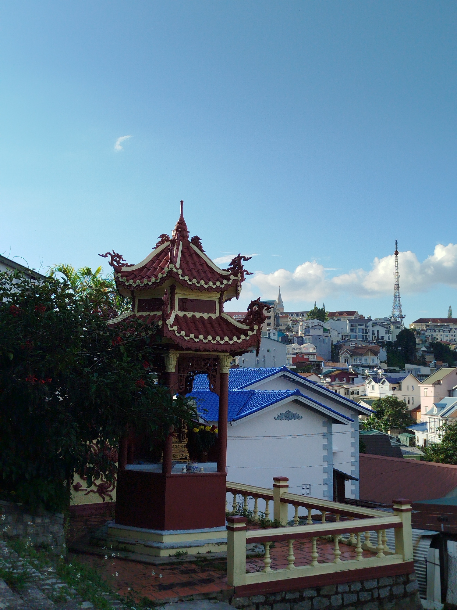 The image is a medium shot of a city landscape in Da Lat, Vietnam, taken from a high vantage point. In the foreground, a red and gold pagoda-style structure stands prominently, partially obscured by lush green foliage and red flowers. To the right of the pagoda, a yellow and red railing lines a paved area.
In the mid-ground, a cluster of buildings with distinctive blue roofs is visible. Beyond that, the city of Da Lat stretches across rolling hills, characterized by a dense arrangement of houses with varied architectural styles and roof colors, including white, gray, and terracotta. A tall, slender radio tower stands out against the skyline. The sky above is a clear, bright blue with a few scattered white clouds, and a faint wispy cloud is visible near the top center. The overall scene suggests a sunny day in a densely populated urban area with a significant amount of greenery.