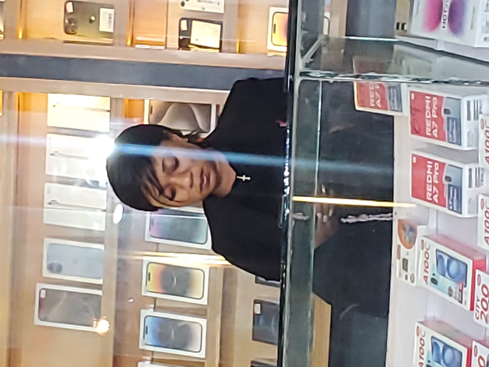 A woman in a black blazer with a cross necklace is standing behind a glass display counter filled with various mobile phones in their packaging. She is looking down at the counter. Behind her, shelves are stocked with more boxed mobile phones, some of which are displayed upright and others lying flat. The environment appears to be a retail store selling electronics. The image quality is low, making it difficult to discern specific details about the woman or the phone models. The location of Jos, Nigeria, is not evident from the visual information in the image.