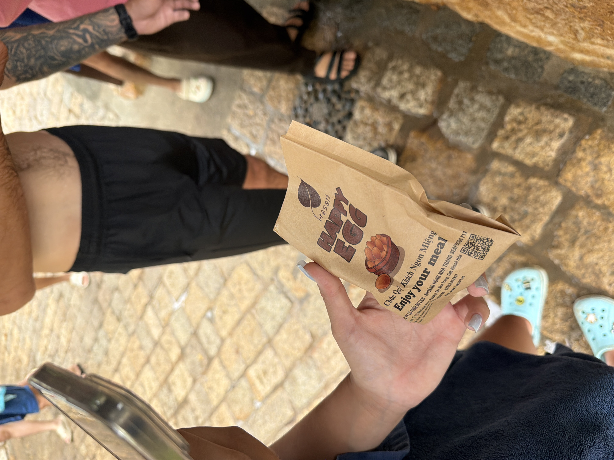 A person's hands are holding a brown paper bag with "Resort Happy Egg" printed on it, along with Vietnamese text. The bag is open, and a small image of eggs in a bowl is visible. The background shows a cobblestone ground and the lower legs of several people wearing casual footwear. The overall impression is that this is an outdoor scene, possibly a market or tourist area in Nha Trang, Vietnam, given the Vietnamese text on the bag. The ground appears wet in places, suggesting recent rain or cleaning.