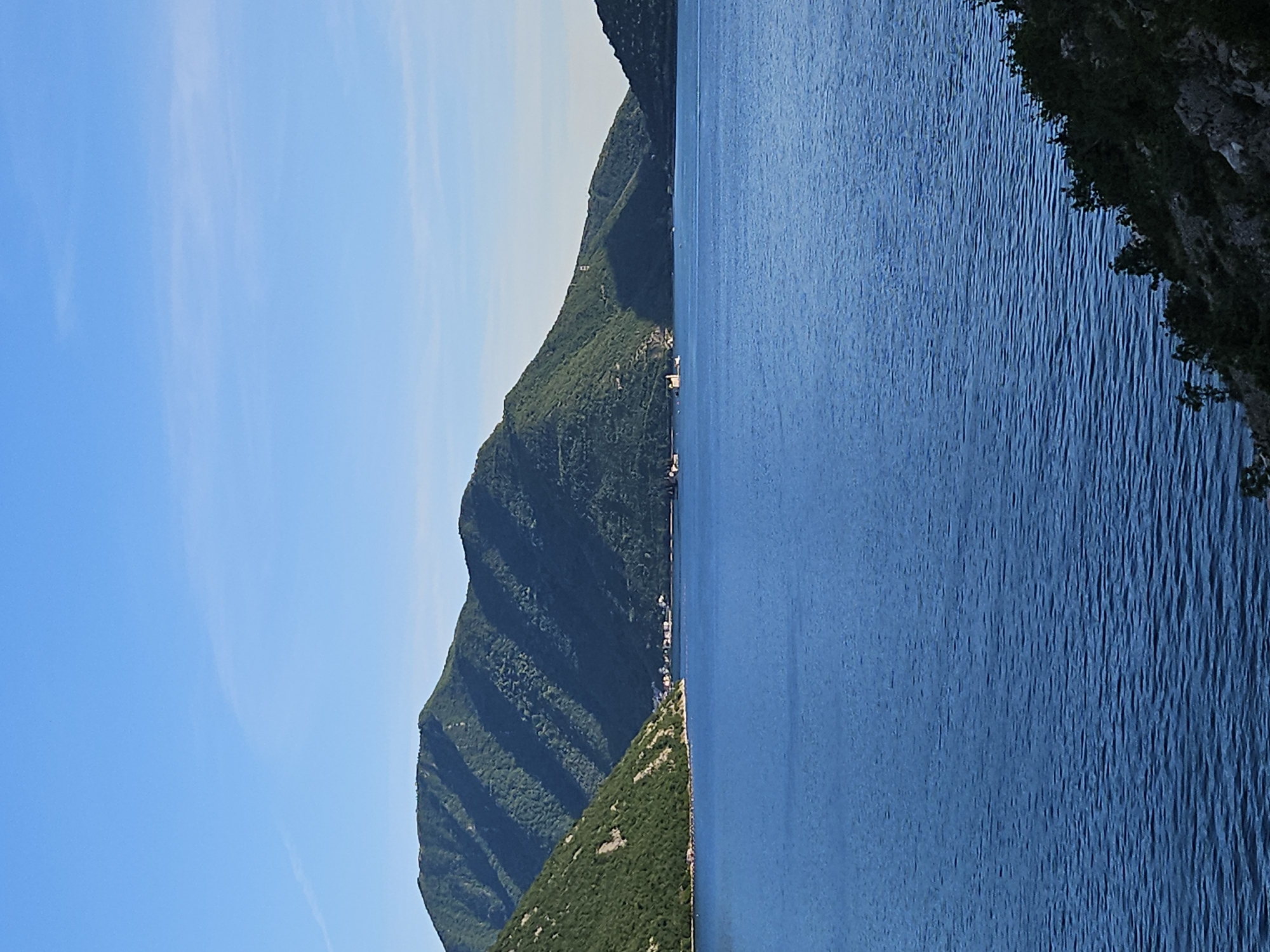 The image shows a landscape of a body of water meeting a mountain range. The sky is a clear blue with a few wispy clouds. The mountains are covered in lush green vegetation. A small town is visible along the shoreline at the base of the mountains. The water is a deep blue color with subtle ripples on the surface. The location is Unknown, Unknown.