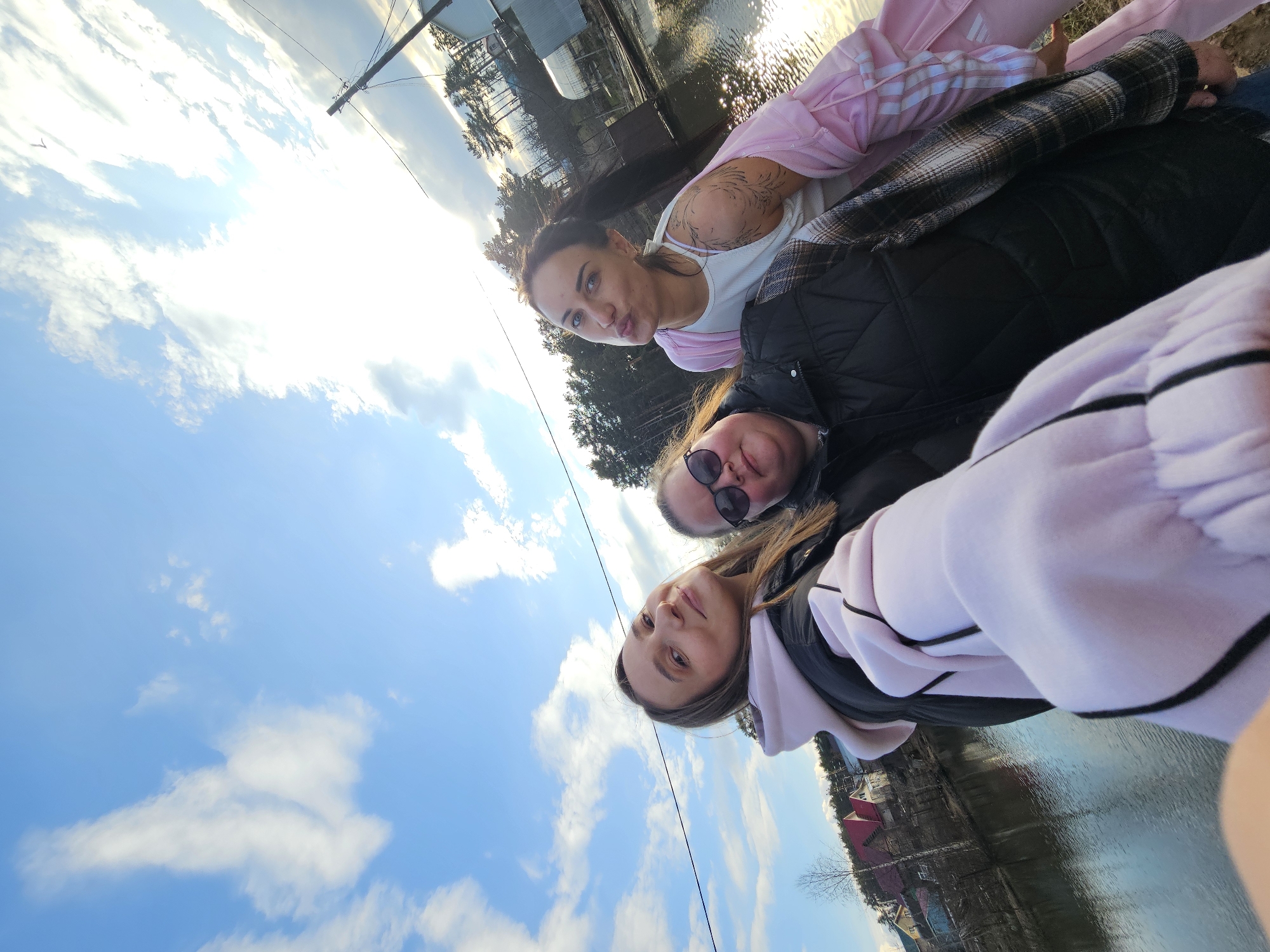 Three women are posing for a selfie. The background shows a river with buildings on the opposite bank. The location is likely in Chaikovsky, Russia. The sky is blue with scattered clouds. One woman is wearing a pink tracksuit, another a black jacket with sunglasses, and the third a light purple sweatshirt.
