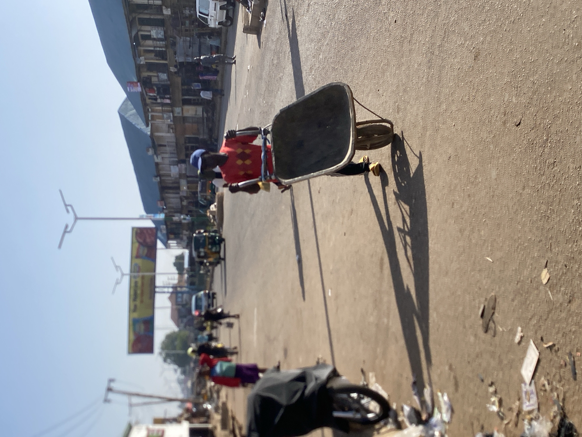 The image depicts a street scene in Bukuru, Nigeria, on a sunny day. A man wearing a red and orange patterned shirt and a blue hat is pushing an empty wheelbarrow along the street. Several other people, motorcycles, and vehicles are visible in the background. Street vendors are present, and a large billboard stands in the distance. The street surface is paved but has debris and trash scattered along the edge. Buildings are on the left side of the image.