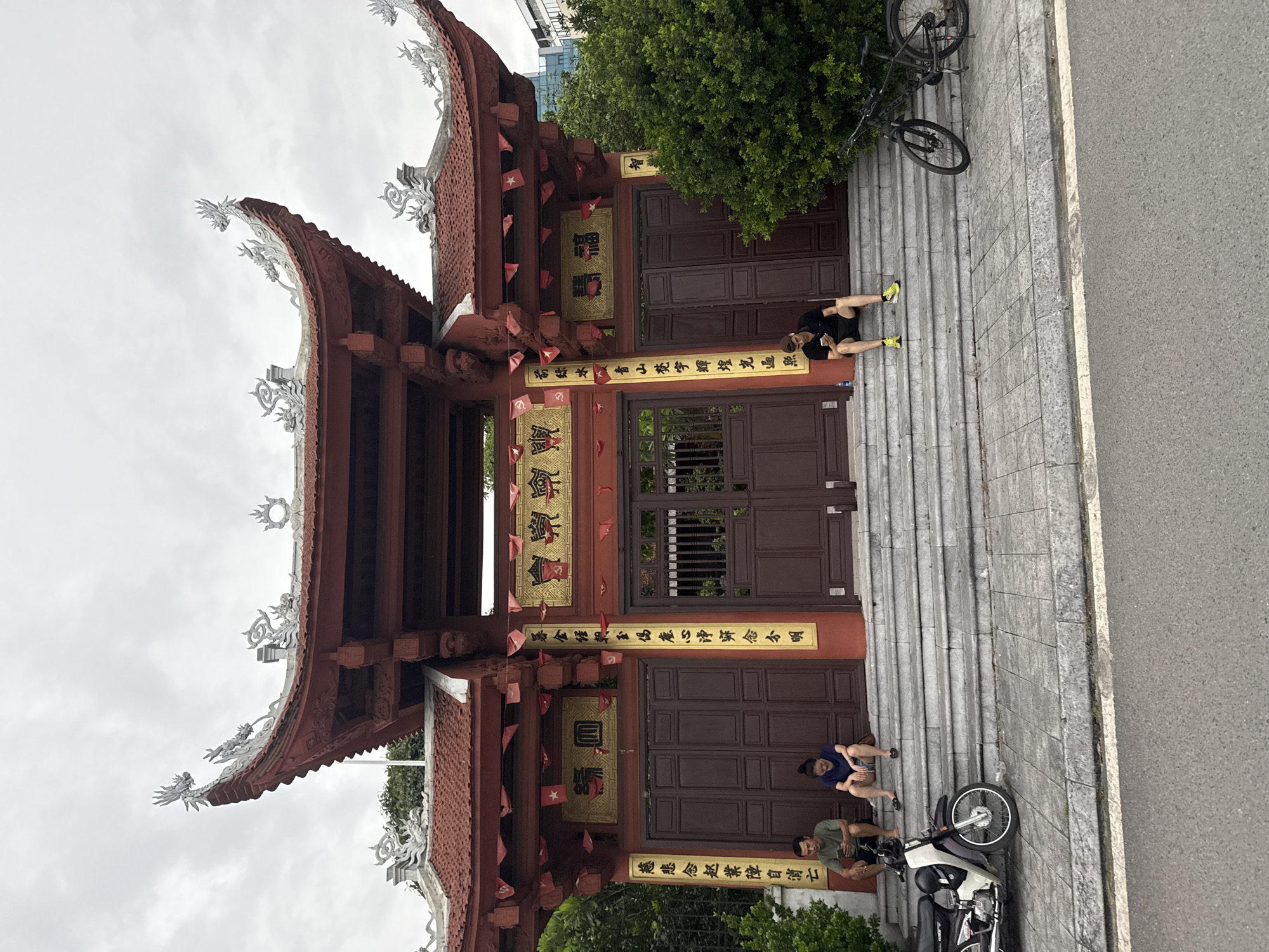 The image shows the entrance to a temple in Hanoi, Vietnam, on an overcast day. The temple features a red, ornate roof with dragon decorations and intricate carvings. A large set of brown wooden doors is centered in the structure, flanked by vertical gold plaques with Vietnamese calligraphy. Small red flags are strung across the top of the entrance.
In front of the temple, a set of stone stairs leads up to the entrance. Three individuals are visible on the stairs. On the left, a man in a green shirt and dark shorts sits looking at his phone, next to a woman in a blue shirt and denim shorts. Further to the right, a man in a black polo shirt and black shorts sits on the stairs looking at his phone.
To the left of the stairs, a white and black motorcycle is parked. To the right of the stairs, a black bicycle is parked. Lush green trees and bushes are visible on the right side of the image, and a portion of a modern building can be seen in the background. The scene is set on a street with asphalt paving in the foreground.