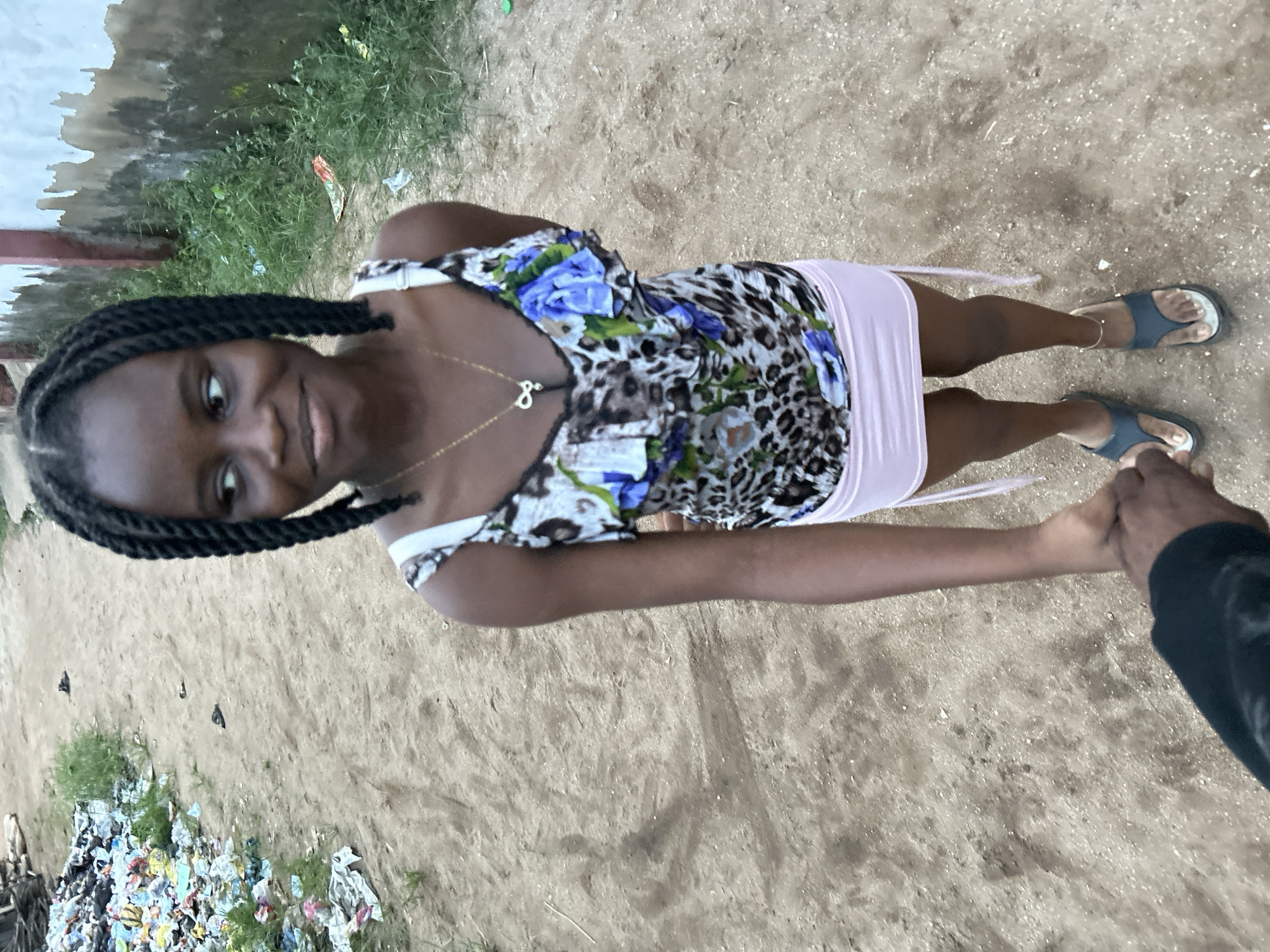 This vertically oriented image, captured from a high angle, features a young Black woman holding hands with another individual in an outdoor setting in Ihiagwa, Nigeria. The woman is looking upwards towards the camera or her companion, displaying a soft smile. She has braided hair and is attired in a strappy top that combines leopard print with vibrant blue floral designs, along with a light pink mini-skirt. She wears dark blue/grey sandals and a necklace featuring a pendant resembling the letter 'B' or the number '8'. Of the second person, only a hand and forearm, clad in a dark long-sleeved garment, are visible, firmly grasping the woman's right hand.
The environment is characterized by a sandy or dirt ground. The background includes a light-colored wall, showing signs of weathering and discoloration, alongside sparse patches of green grass and weeds. Prominently visible in the lower left portion of the image is a substantial accumulation of litter, including plastic bags and other refuse, scattered across the ground. The lighting appears soft and diffused, suggesting it is daytime, possibly under an overcast sky or during the late afternoon, as there are no harsh shadows. The intimate gesture of hand-holding and the woman's direct, warm gaze convey a sense of connection and contentment.