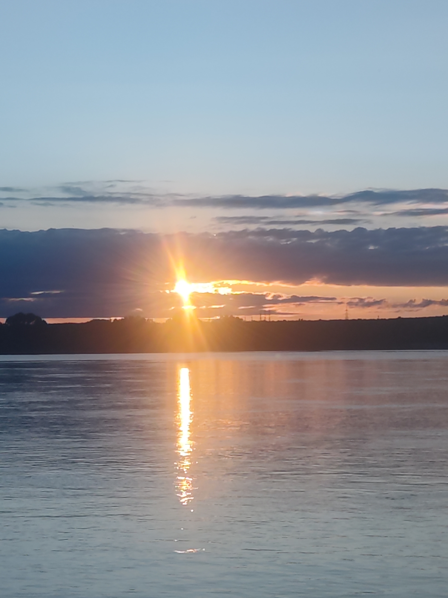 The image presents a serene sunset over an expansive body of water, likely a river or reservoir near Chaikovsky, Russia. The sun, a brilliant golden disk, is positioned near the center of the frame, partially obscured by dark, horizontal cloud banks along the horizon. Its intense rays burst forth, fanning across the sky and creating a spectacular display of light.
The sky transitions from a clear, pale blue at the top to darker blues and grays near the horizon, where more substantial cloud formations enhance the dramatic effect of the setting sun. These clouds filter the sunlight, contributing to the warm hues visible in the lower sky.
A dark, silhouetted landmass stretches across the horizon, appearing as a distant shore or hilly terrain. Within this silhouette, faint suggestions of structures, possibly power line towers, can be discerned, indicating some human presence in the remote landscape.
The calm surface of the water dominates the foreground and midground, acting as a mirror for the sun's radiant light. A vivid, shimmering path of golden and orange reflections extends directly from the sun towards the bottom of the image, dancing on the subtle ripples across the water.
The scene captures a moment of natural beauty and tranquility, evoking feelings of peace and awe. The time of day is clearly sunset, characterized by the warm, fading light. The weather appears partially cloudy, which enhances the visual drama of the light. There are no people or discernible activities taking place, and no visible text.