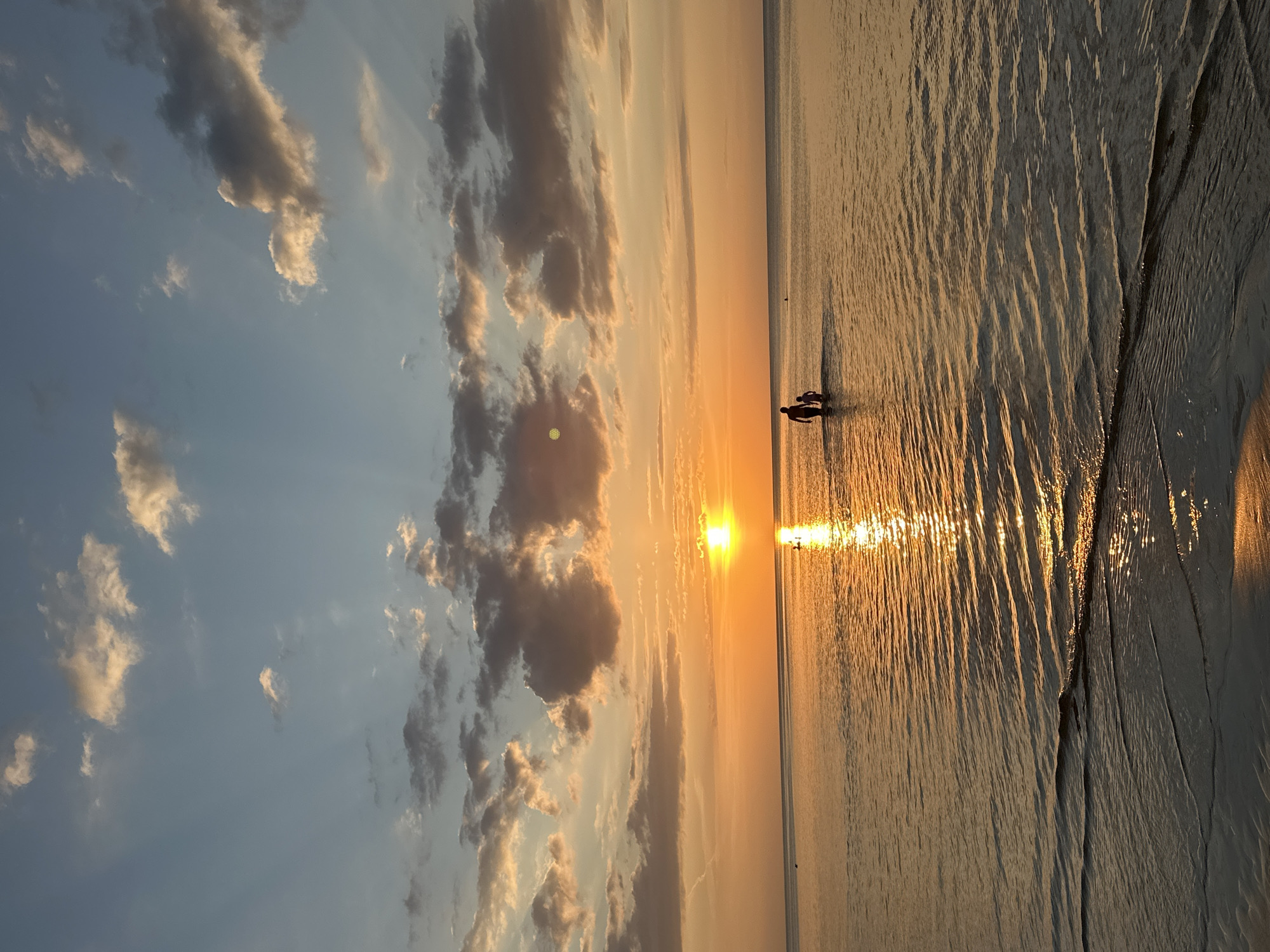 A sunset over the Baltic Sea in Jūrmala, Latvia. Two silhouetted figures, an adult and a child, are walking through the shallow water towards the horizon. The sun is low in the sky, casting a warm, golden glow across the water and reflecting off its rippling surface. The sky above is a mix of pale blue and soft orange, with scattered clouds tinged with pink and gold. The foreground shows the edge of a sandy beach, wet from the receding tide.
