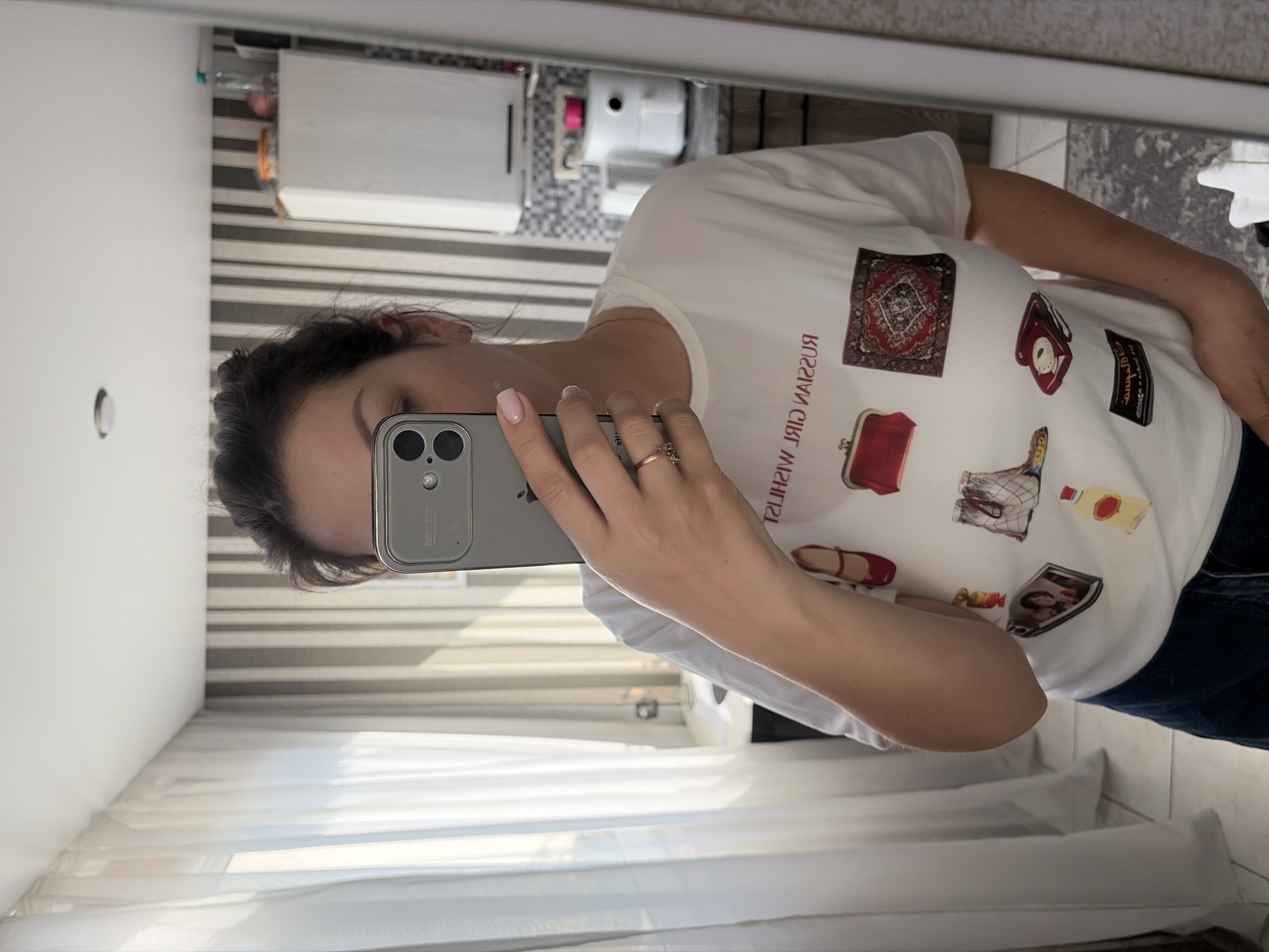 A woman, seen from the chest up, stands in front of a mirror and takes a selfie with her iPhone. She is wearing a white t-shirt adorned with various graphics. The graphics include a red purse, a patterned rug, a pair of red high-heeled shoes, a pair of fishnet stockings, a red rotary phone, a framed photograph, and a bottle of yellow liquid. The woman has dark hair pulled back in a bun and is wearing a ring on her right ring finger. Her nails are painted pink.
The environment appears to be an indoor space, likely a bathroom or dressing area. Behind her, vertical striped wallpaper is visible, along with a white cabinet and what looks like a kitchen appliance. To her left, white sheer curtains hang, suggesting a window is present. The overall impression is a casual, personal snapshot taken in a domestic setting. There are no elements in the image that specifically indicate a location in Sochi, Russia.
