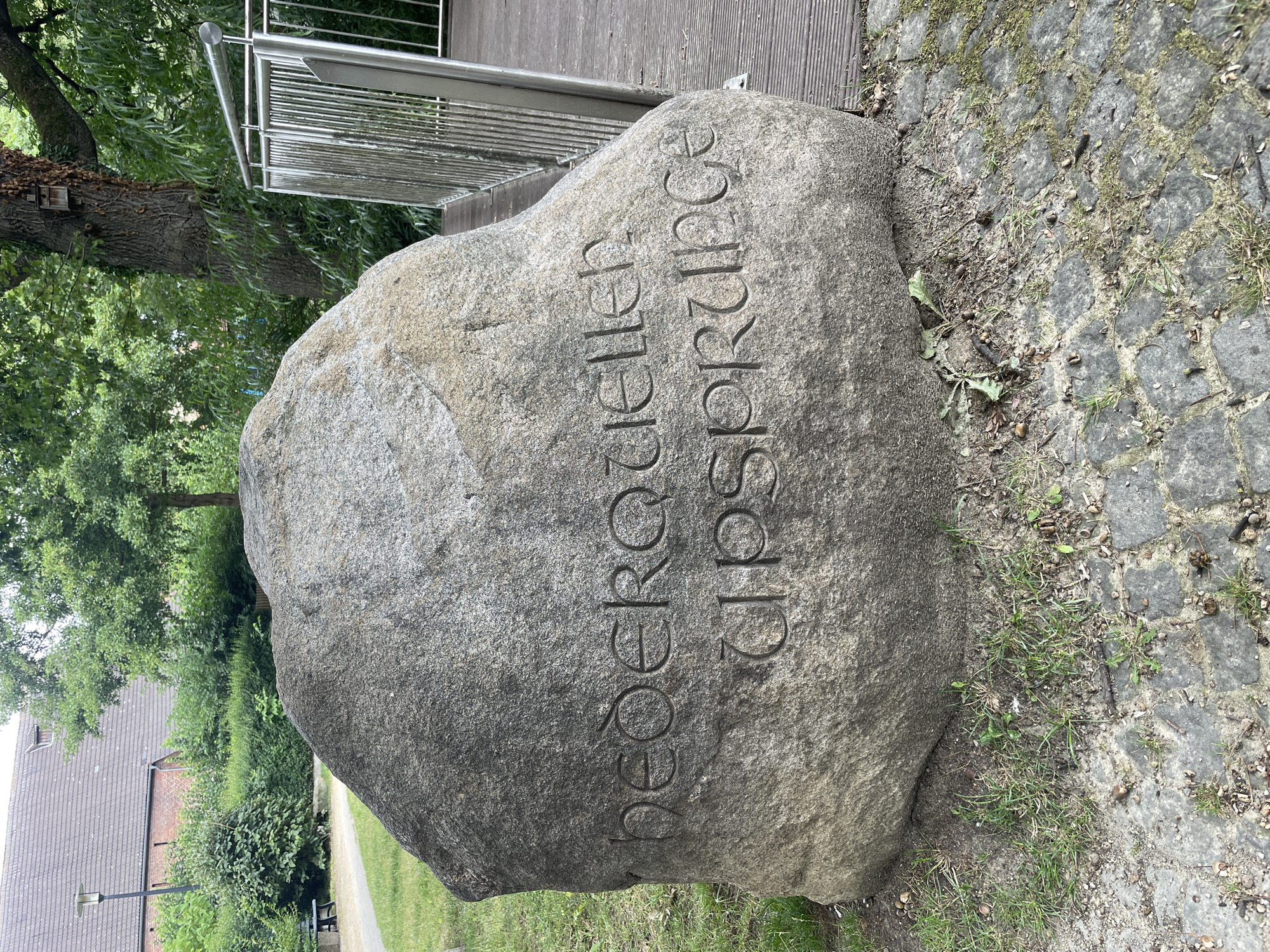 A large, rough-hewn grey boulder is the primary subject, positioned outdoors on a grassy area with cobblestone paving visible in the foreground. The boulder has carved lettering on its surface. The top line of text reads "HEDERQUELLEN" and the bottom line reads "URSPRÜNGE". Behind the boulder, to the left, a building with a red-tiled roof and a dormer window is visible, surrounded by green trees and shrubs. A lamppost stands near the building. To the right of the boulder, a wooden walkway with a silver metal railing is present, leading towards the background where more trees and foliage are seen. The lighting suggests it is daytime, with diffused sunlight. The location is the historic town of Salzkotten, Germany, specifically at the source of the river "Heder".