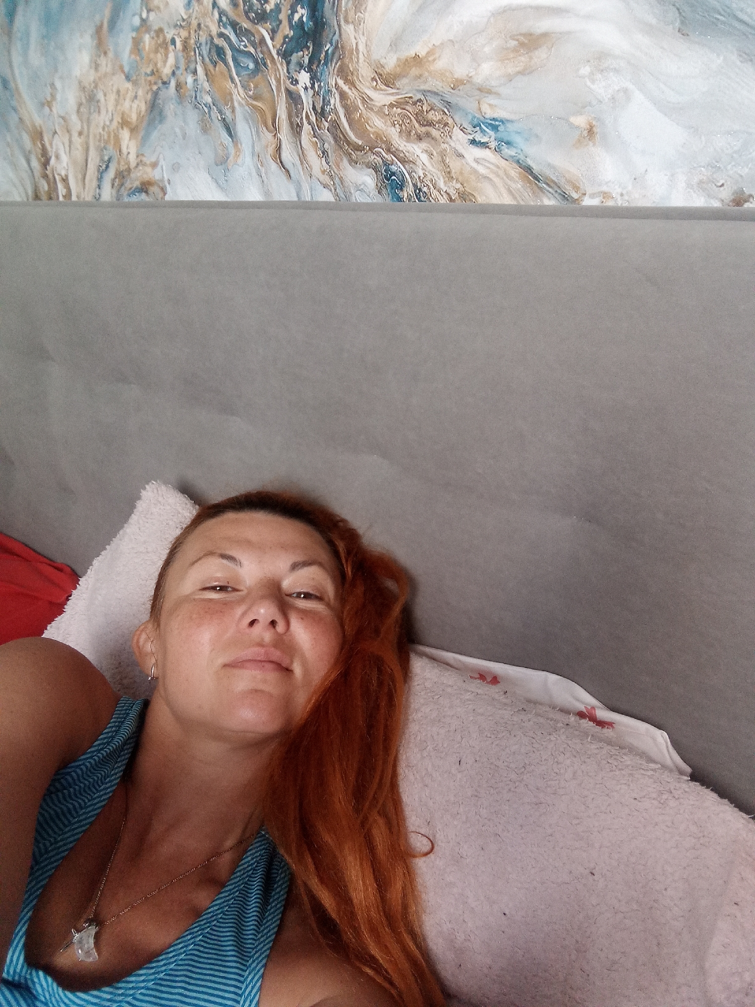 A woman with auburn hair lies on her side on a pillow, looking at the camera. She is wearing a blue and white striped tank top and a silver pendant necklace. Her right arm is visible, resting on the pillow. The background is a gray wall with a piece of abstract art above it. The lighting is soft and natural, casting a gentle glow on the woman's face.