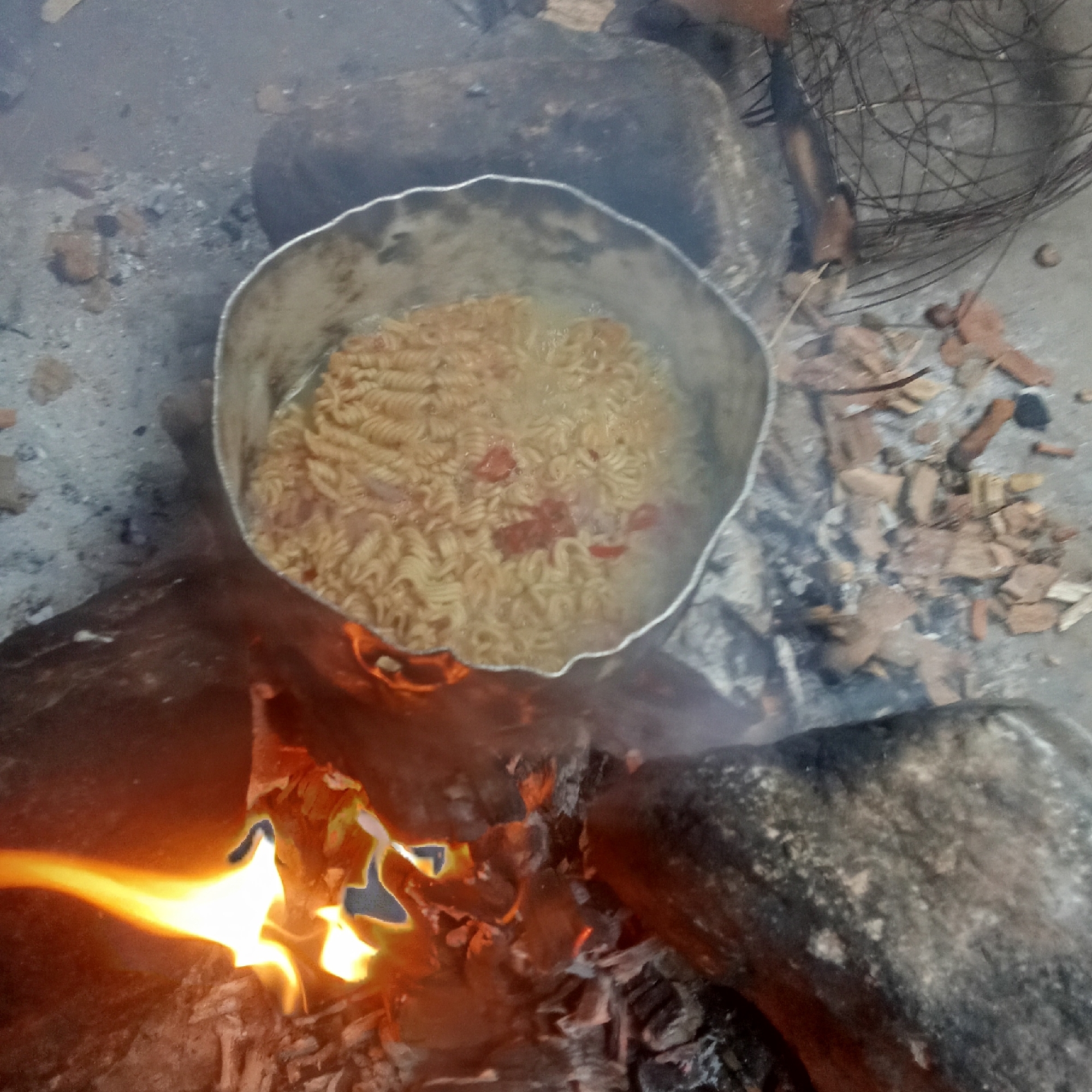 A metal pot containing noodles and some tomato pieces sits atop a wood fire, which is contained within a rough circle of large stones. The scene is set outdoors, on a patch of ground littered with ash, bits of wood, and small brown debris. A wire basket lies to the upper right. The location is Unknown, Unknown.