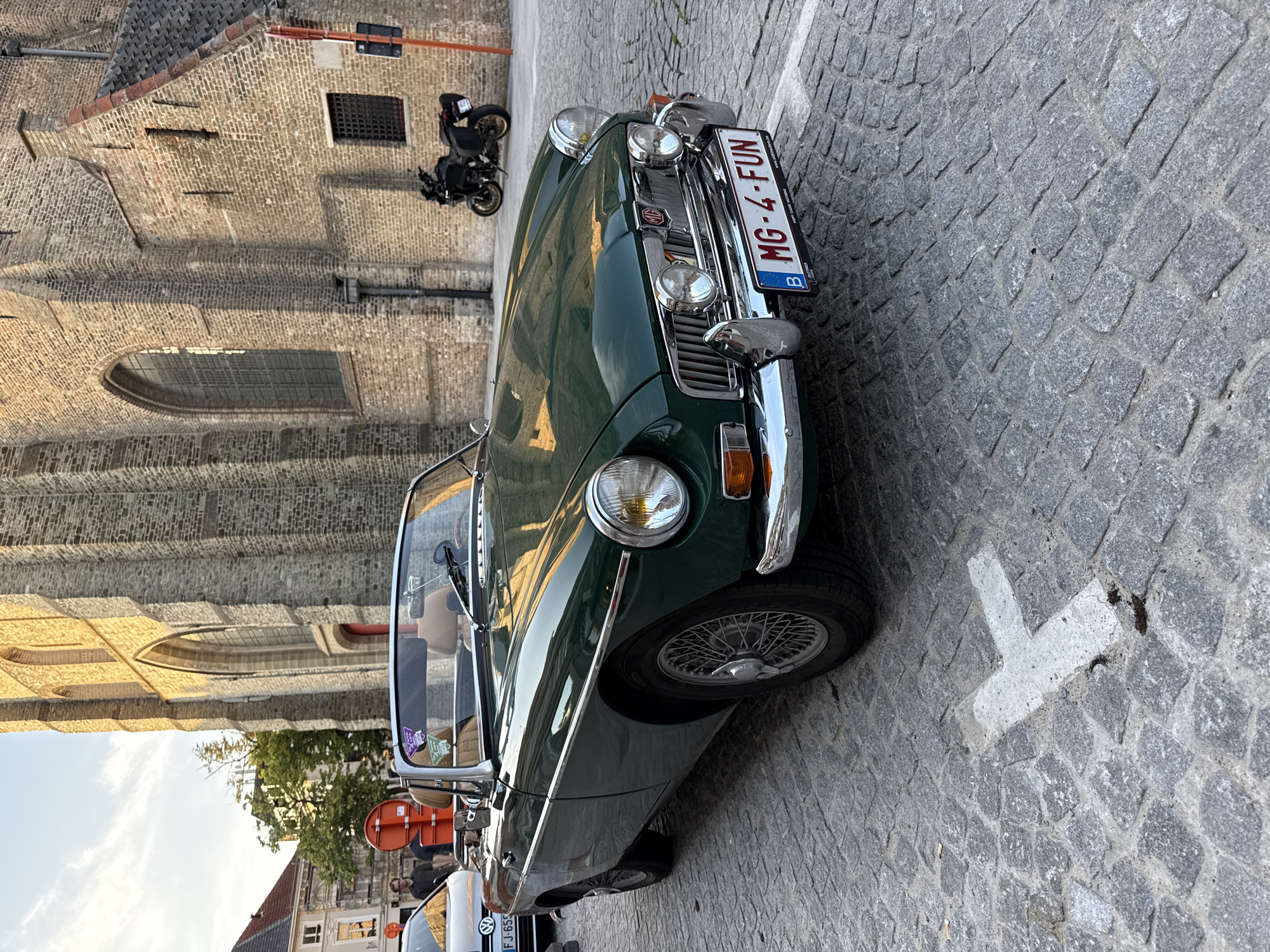 This is a medium-shot, eye-level, outdoor photograph of a classic dark green convertible car parked on a cobblestone street. The car is the primary subject, with its front grille, headlights, and license plate prominently displayed. The license plate reads "MG-4-FUN" and appears to be a Belgian plate, indicated by the letter "B". The car has chrome accents and wire-spoke wheels.
Behind the car, a weathered brick building with arched windows and a tiled roof is visible. The building's texture suggests it is old, possibly a church or historical structure, consistent with the location context of Bruges, Belgium. To the left of the car, a black motorcycle is parked near the building. The street is paved with grey cobblestones, and a white painted cross shape is visible on the ground in the foreground, likely a road marking.
The lighting suggests it is daytime, with shadows indicating sunlight. The overall mood is calm and pleasant, capturing a scenic moment in a historic European city. There are no people prominently featured in the foreground, but a reflection in the car window suggests at least one person may be present. The image is oriented vertically.