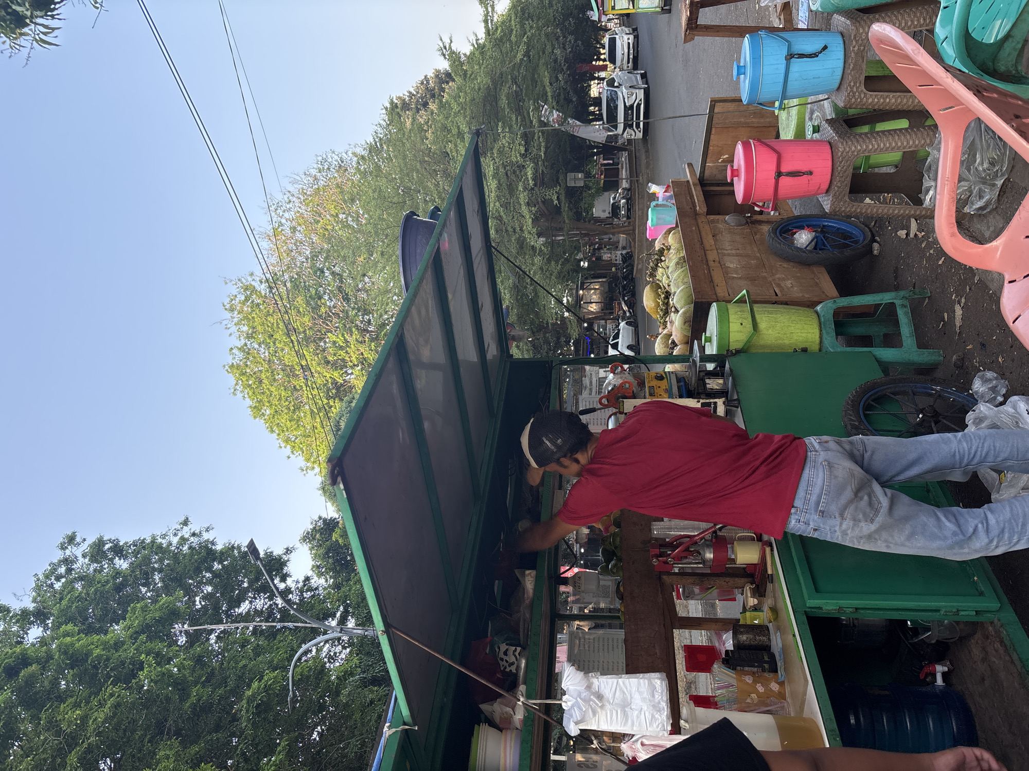 An outdoor scene in Cirebon, Indonesia, features a street vendor's cart laden with coconuts and other produce. A man in a red shirt and blue jeans is working at the cart, arranging items under a green awning. Several plastic containers in bright colors – pink, blue, and green – are placed on small stools, along with some plastic chairs. In the background, a street with cars and trees suggests a moderately busy urban environment. The overall impression is of a typical street-side vendor stall in Indonesia.