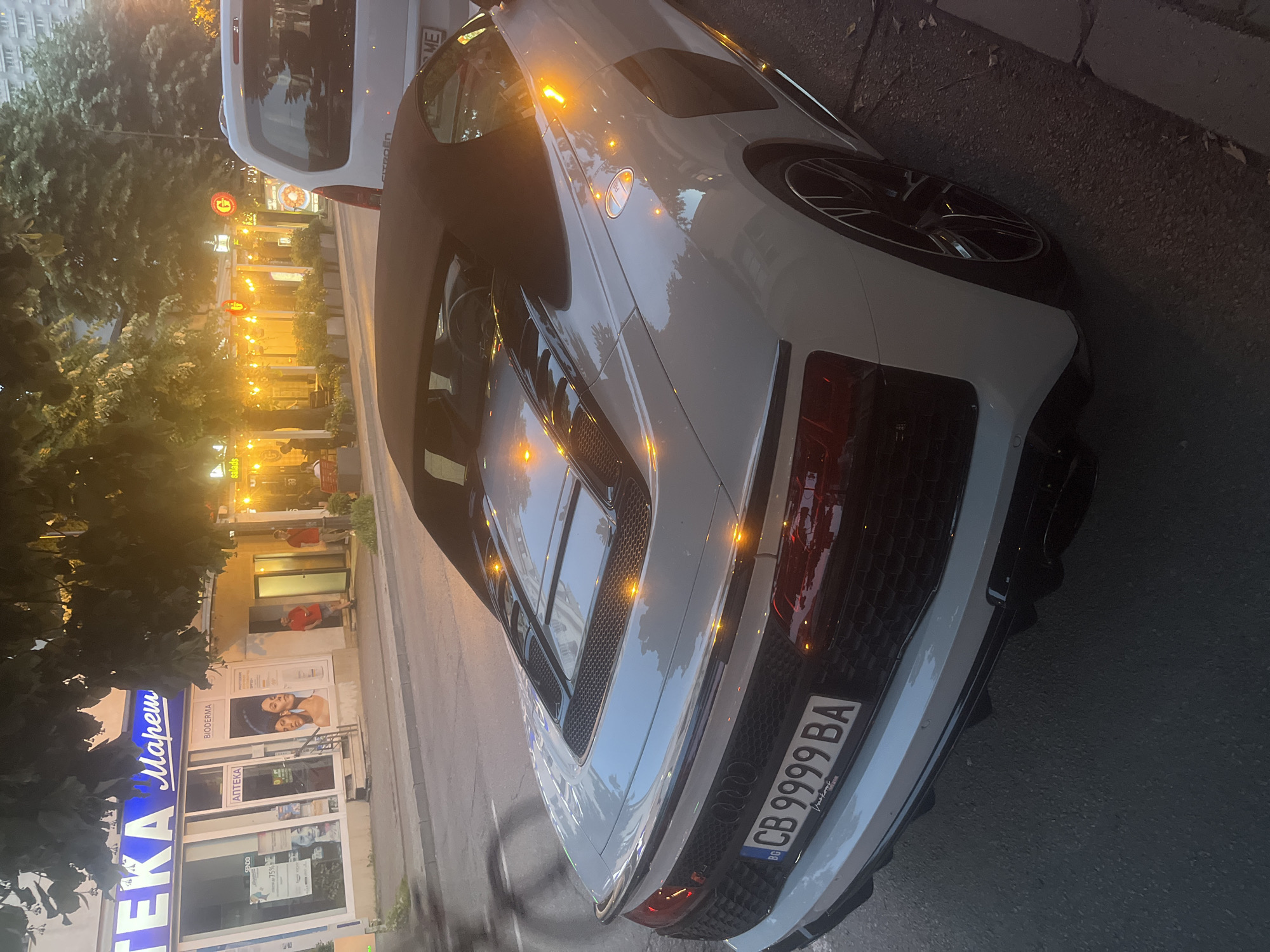 A white convertible Audi R8 is parked on a street in Varna, Bulgaria, at dusk. The car is facing away from the camera, with its rear end prominently displayed. The license plate reads "CB 9999 BA". In the background, there are storefronts with illuminated signs, including "АПТЕКА МАРЕШ" and "BIODERMA". A white van is parked behind the Audi. People are visible in front of the storefronts. Trees and streetlights also contribute to the urban nighttime scene.