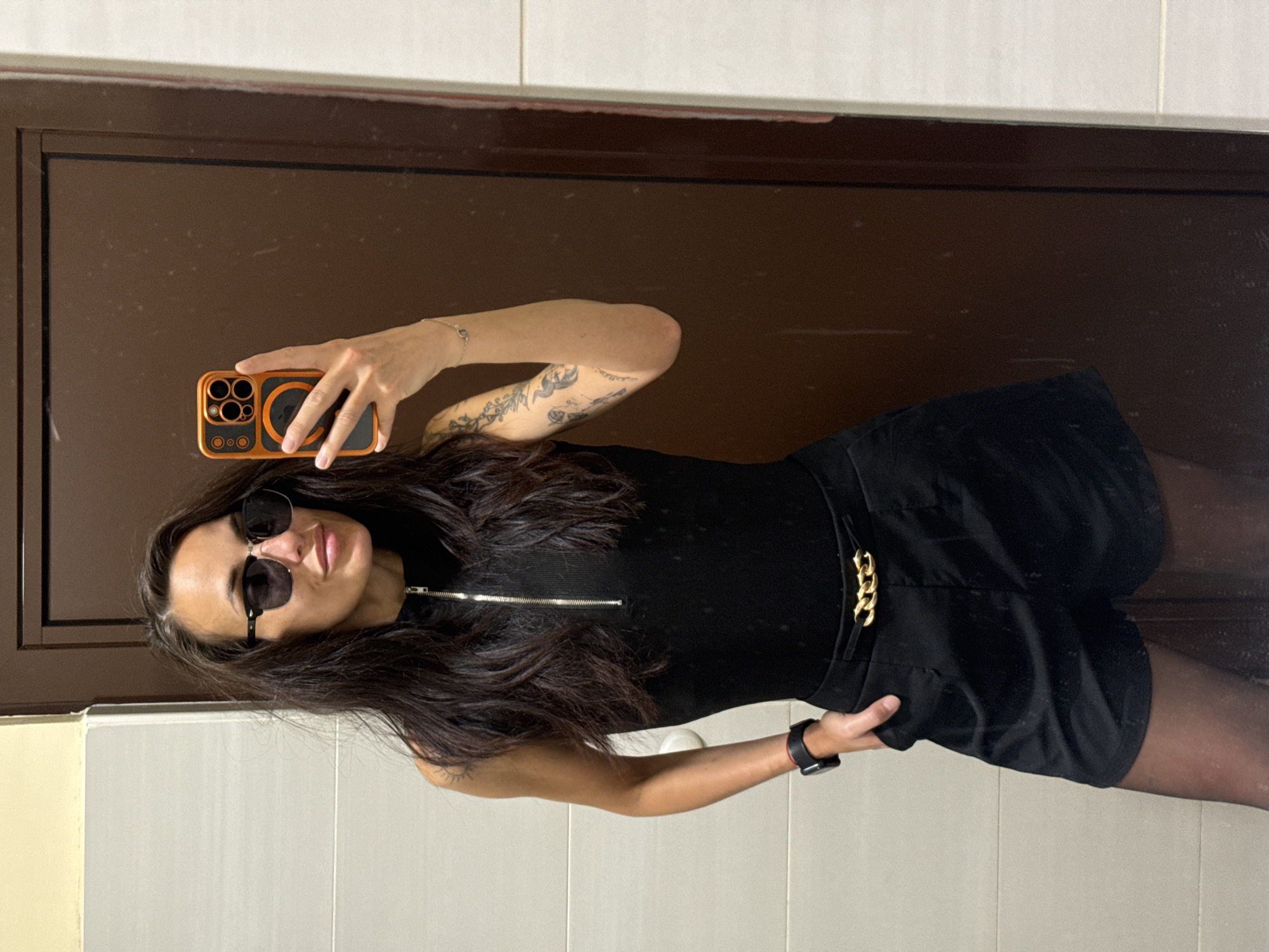 A woman stands indoors, likely in a bathroom setting, taking a selfie in a mirror. She is wearing a black outfit, consisting of a top with a zipper detail and black shorts with a gold chain accent. She has black tights on her legs and is wearing sunglasses. In her left hand, she holds an orange smartphone, while her right hand is positioned on her waist. Her dark hair is long and wavy. The background shows a brown door with a light-colored tiled wall. The image does not provide specific information to identify the location as Byala Slatina, Bulgaria.