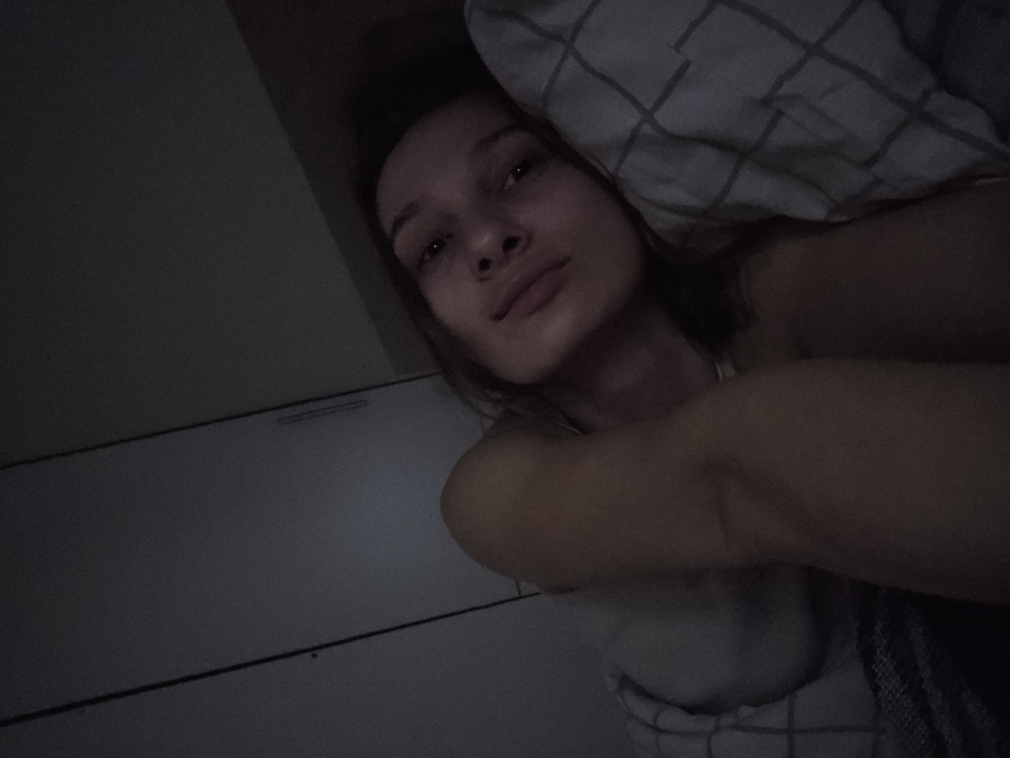 A woman is lying in bed with her head resting on a pillow that has a grey and white checkered pattern. She is looking at the camera with a neutral expression. Her left arm is raised and partially obscures her chest. The room is dimly lit. To the left of the woman is a wall with a built-in cabinet that has horizontal lines. The location is likely a bedroom but cannot be confirmed to be in the city of Chaikovsky, Russia based on the given information.