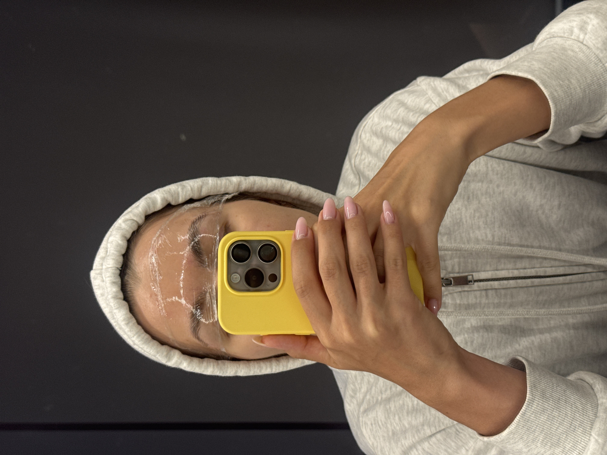 A person with a clear plastic wrap over their eyebrows, wearing a grey hoodie, is taking a selfie with a yellow phone. The person's face is partially visible, and they have manicured nails. The background is dark and indistinct. The location is not specified.