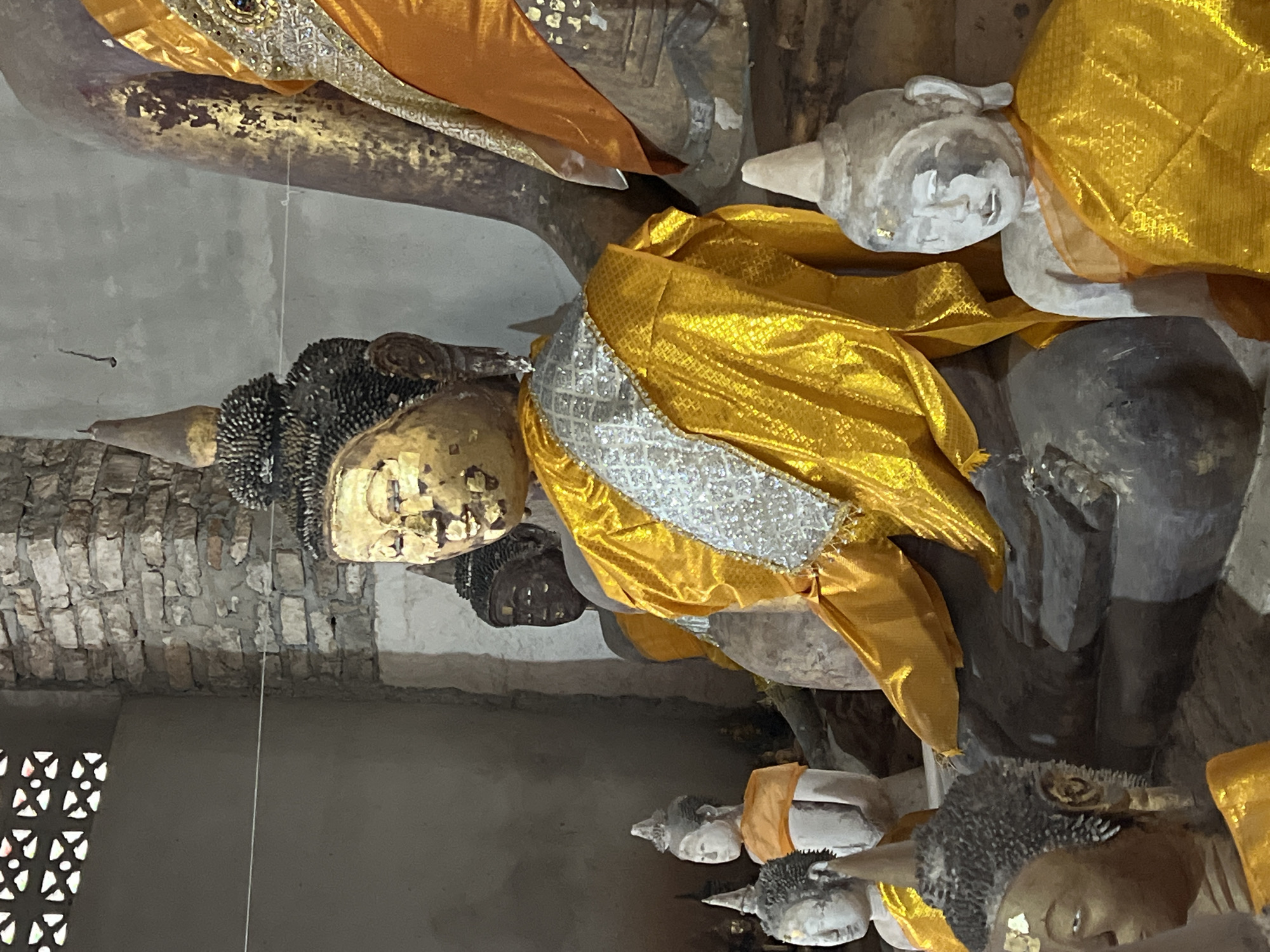 The image displays a collection of Buddha statues within an indoor environment, likely a temple or shrine in Ko Samui District, Thailand. The statues vary in size and material, with some appearing to be made of stone and others of a darker, possibly bronze or wood material. Several statues are adorned with vibrant yellow and orange robes, some of which are intricately patterned. One prominent statue in the foreground, with a gold-painted face, wears a yellow robe with a decorative silver band across its chest. The background reveals a rough brick wall and a portion of a large, darker statue, also draped in fabric. The lighting is subdued, suggesting an interior setting, and the overall atmosphere is one of reverence and age.