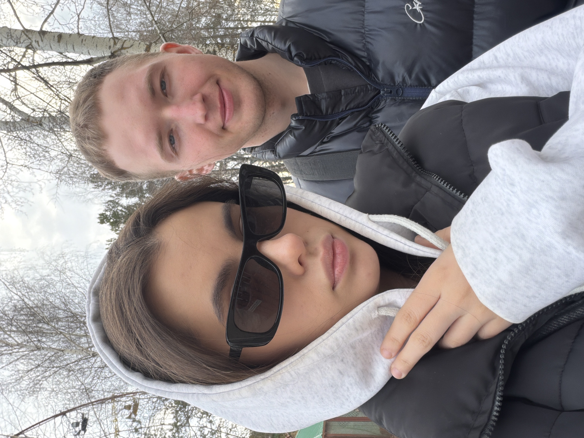 This is a vertical selfie featuring two individuals, a male and a female, captured outdoors in what appears to be a natural, possibly park-like setting in Chaikovsky, Russia.
The female subject is in the foreground, wearing a light grey hooded sweatshirt and a black puffer jacket or vest. She has long, dark brown hair, some of which is visible beneath her hood. She is also wearing dark, rectangular sunglasses that obscure her eyes, and her lips are slightly parted. Her right hand is visible, resting on her jacket.
Behind and slightly above her is the male subject. He has short, light brown hair and a subtle, pleasant smile. He is dressed in a dark, likely black or navy, puffer jacket with a visible zipper closure. A small, stylized white emblem resembling a 'C' or similar symbol is embroidered on the upper left chest of his jacket.
The background consists of numerous bare trees, characteristic of a cool season such as late autumn, winter, or early spring. Prominently featured are white-barked birch trees. The sky is overcast and bright, indicating daytime. Both individuals' attire suggests protection from cold weather. The overall impression is one of a relaxed, casual moment shared between the two subjects.