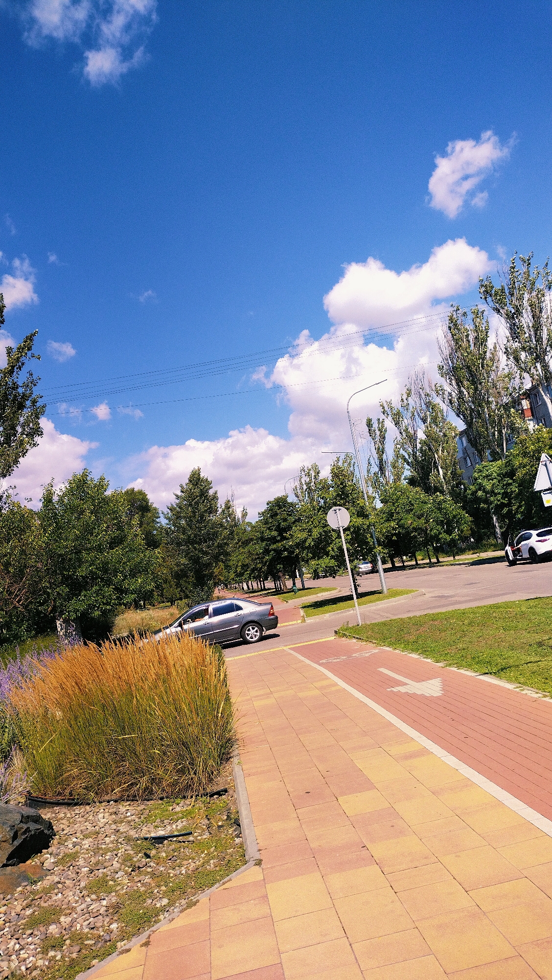 This is a photograph taken on a sunny day in Horishni Plavni, Ukraine, depicting a street scene with a blue sky dotted with white clouds. In the foreground, a pedestrian walkway paved with alternating yellow and red rectangular tiles runs diagonally from the bottom left to the top right. To the left of the walkway, there is a patch of dry, golden grass, some purple flowers, and loose stones. A dark grey sedan is visible driving on the asphalt road adjacent to the walkway. The road is lined with green grass and trees. Further down the road, several tall, slender trees with green foliage are prominent. In the background, apartment buildings can be seen behind the trees. A lamppost stands near the walkway. A white car is parked on the right side of the road.