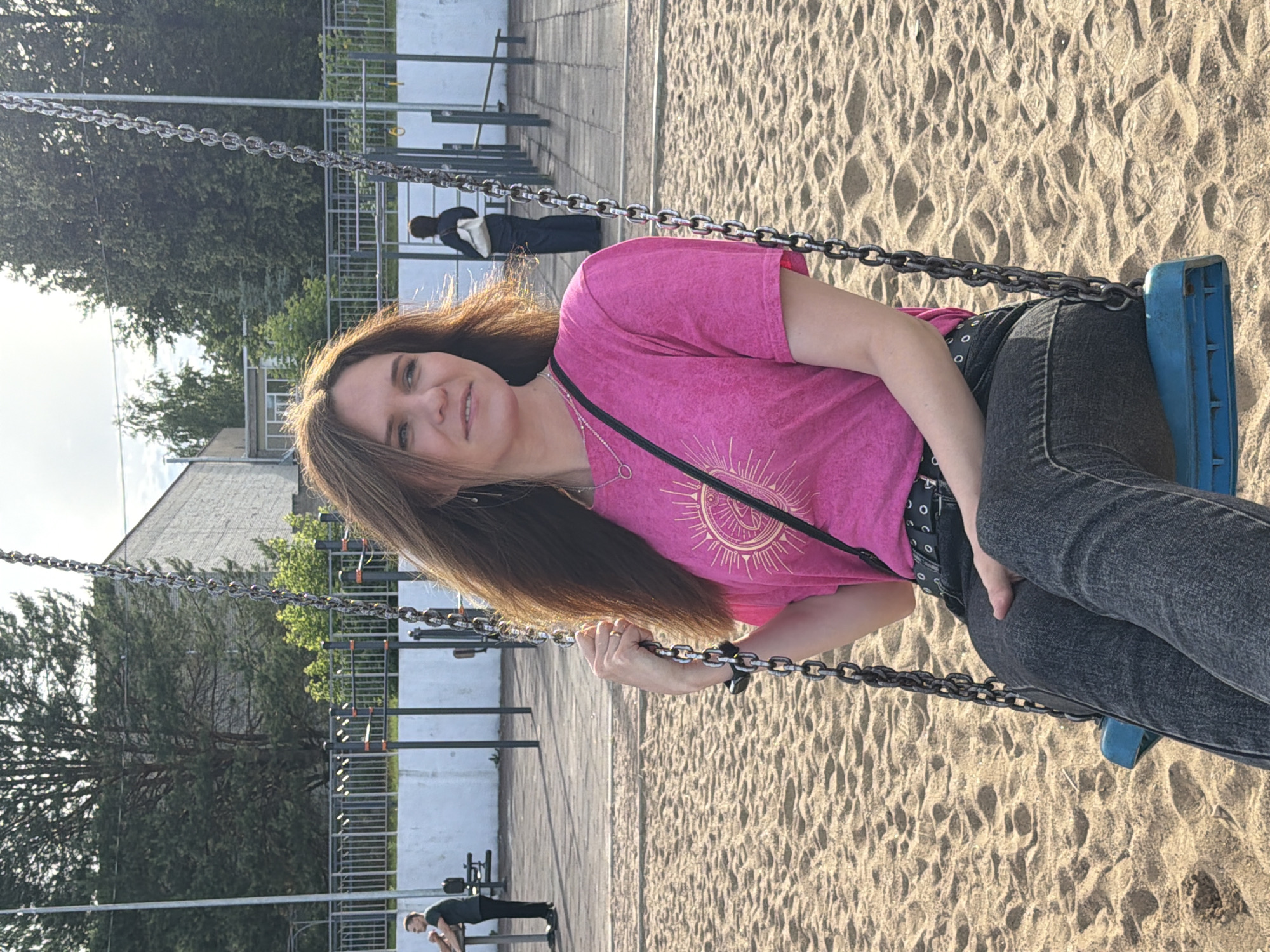 A woman is on a swing set at a playground. She is wearing a pink t-shirt and dark jeans. She is smiling and looking at the camera. The swing set is made of metal chains and a blue seat. The ground beneath the swing set is covered in sand. In the background, there is a building and trees. The sun is shining, indicating it is daytime. Another person is visible in the background, in the lower left corner, in a plank position on a piece of playground equipment. Another figure in a dark coat is visible on a swing further in the background. The scene is set in a park or outdoor recreational area.