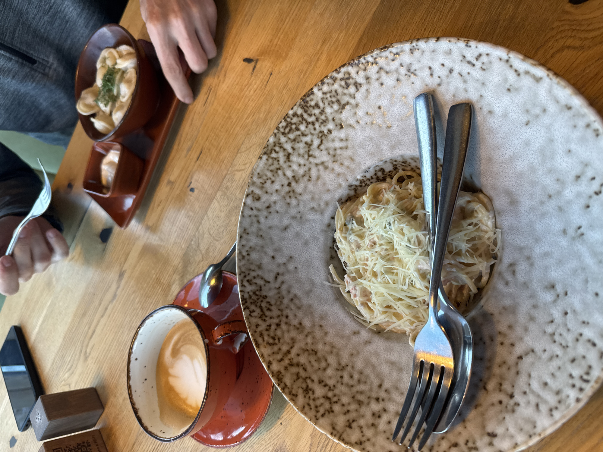 The image shows a close-up of a wooden table at a cafe in Kurkijoki, Russia. On the table, there is a plate of pasta topped with grated cheese, a cup of coffee with latte art, a small dish of sauce, and a bowl of dumplings. A fork and spoon rest on the pasta plate. In the background, a person's hands are visible, one holding a fork and the other reaching towards the dumplings. A smartphone and a salt shaker are also on the table. The cafe appears to have a rustic ambiance with its wooden furniture.