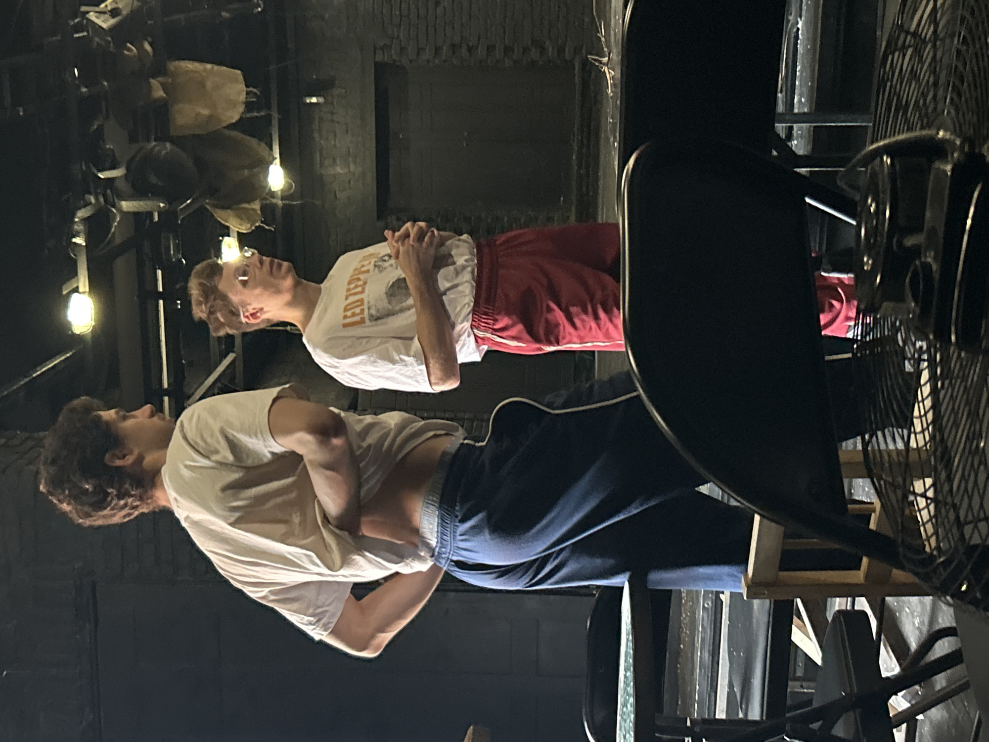 Two young men are standing in what appears to be a theater or rehearsal space. The man on the left, facing away from the camera, is wearing a white t-shirt and dark blue sweatpants. He is pulling the bottom of his t-shirt up, exposing his midriff and a glimpse of his waistline. The man on the right is facing the camera and is wearing a white t-shirt with "LED ZEPPELIN" printed on it and red athletic shorts. He is wearing glasses and has his hands clasped in front of him. The background is dark, with exposed brick walls and theatrical lighting equipment hanging from the ceiling. There are several chairs and tables visible in the foreground, suggesting a backstage or seating area. The lighting is dim, with some spotlights creating bright areas.