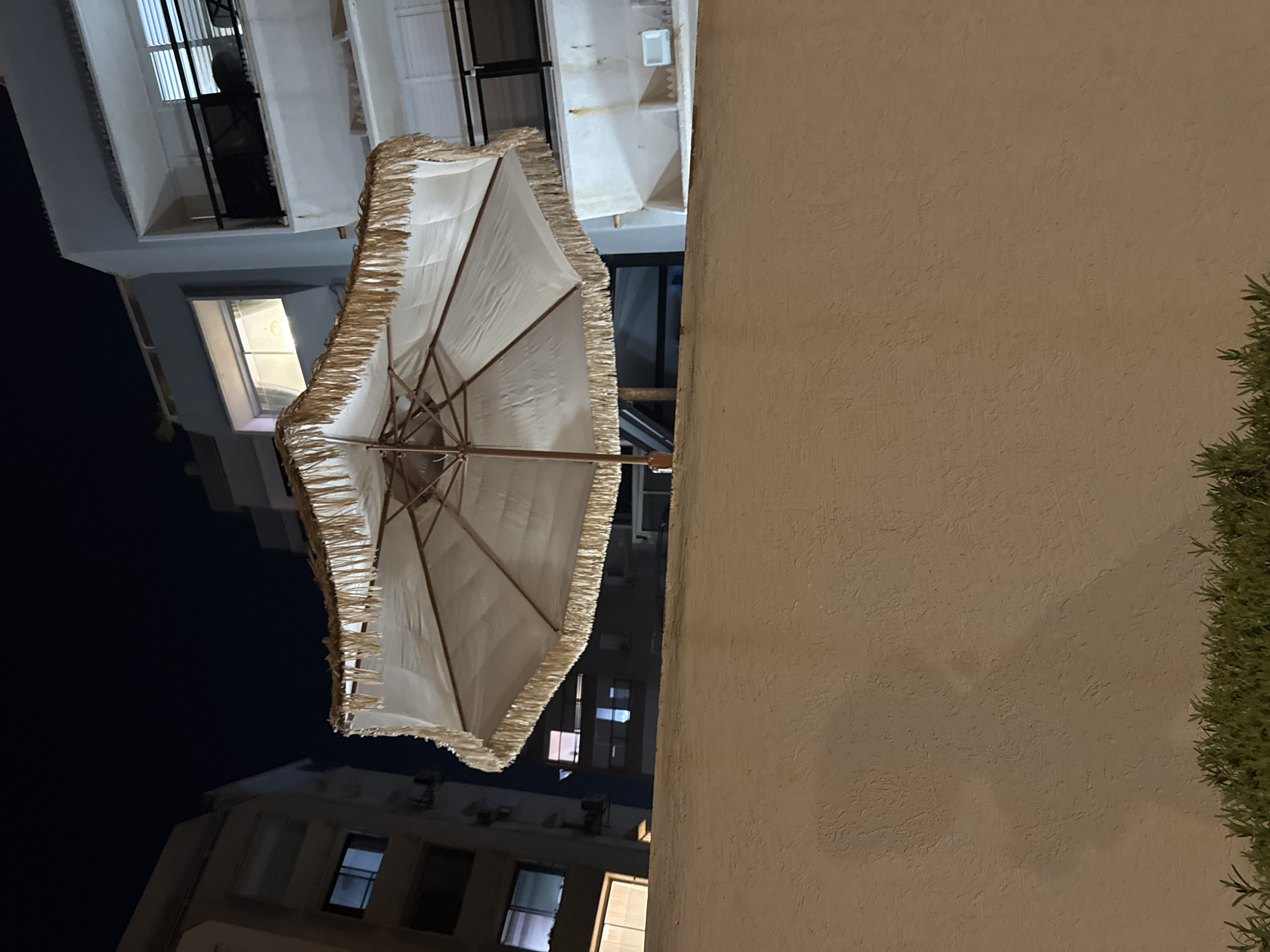 An open, white patio umbrella with a fringed edge is positioned in front of a textured, beige wall at night. The umbrella's central pole is visible, and its canopy is angled upwards. Behind the wall, buildings with illuminated windows are visible, suggesting an urban environment. The scene is likely in Netanya, Israel, as indicated by the context. The overall lighting suggests it is nighttime. There are no people or visible actions occurring in the image.