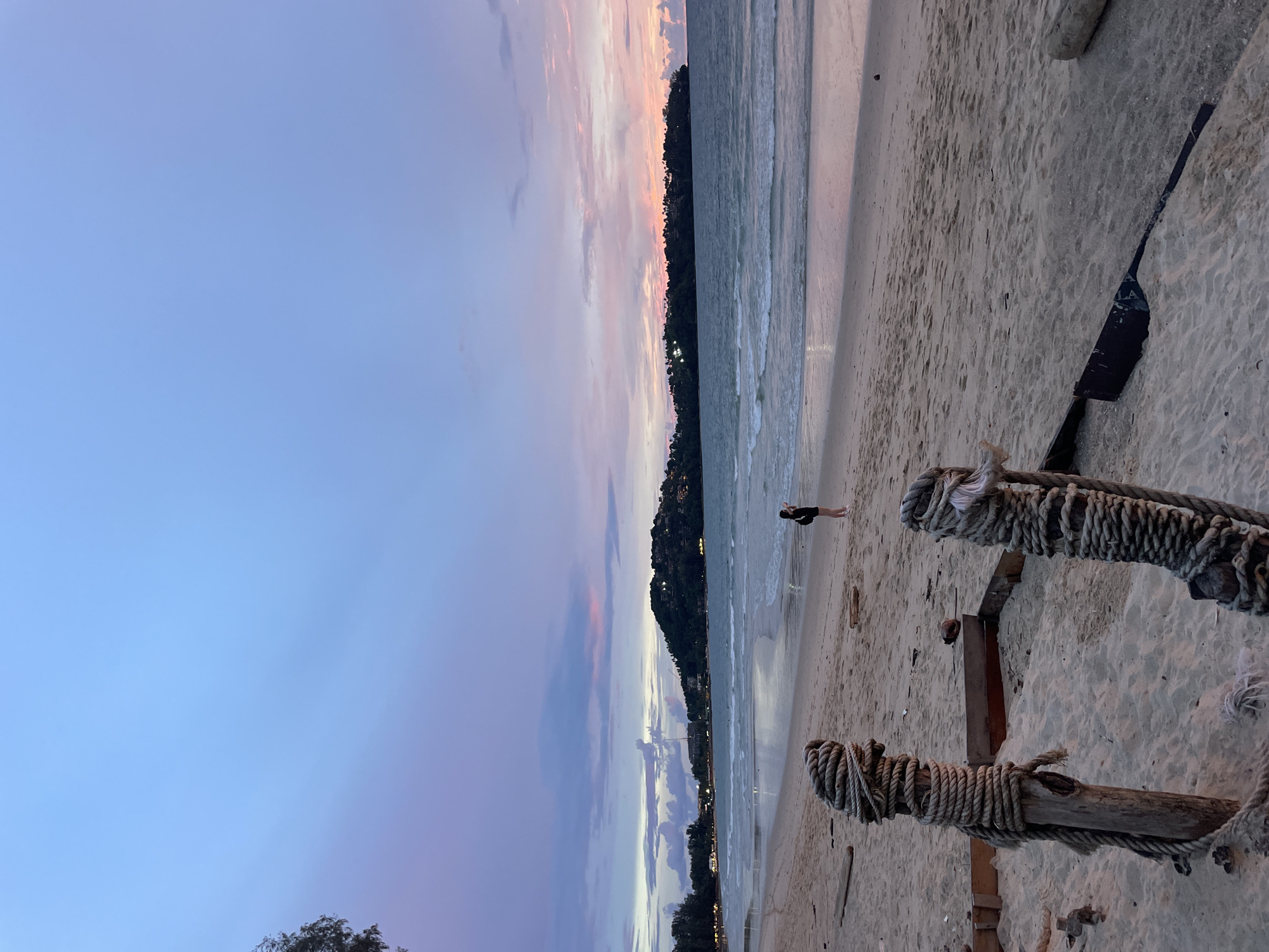 A person is walking on a sandy beach at dusk. The sky is a gradient of blues, purples, and pinks, indicating sunset or twilight. The ocean has gentle waves, and the water is a muted blue-gray. In the foreground, two wooden posts wrapped with thick rope stand on the sand. A dark object with the word "LA" is partially visible in the sand near the right side of the image. The background features a tree-lined hillside with some lights suggesting buildings. The overall mood is peaceful and serene. The image appears to be taken in Choeng Thale, Thailand.