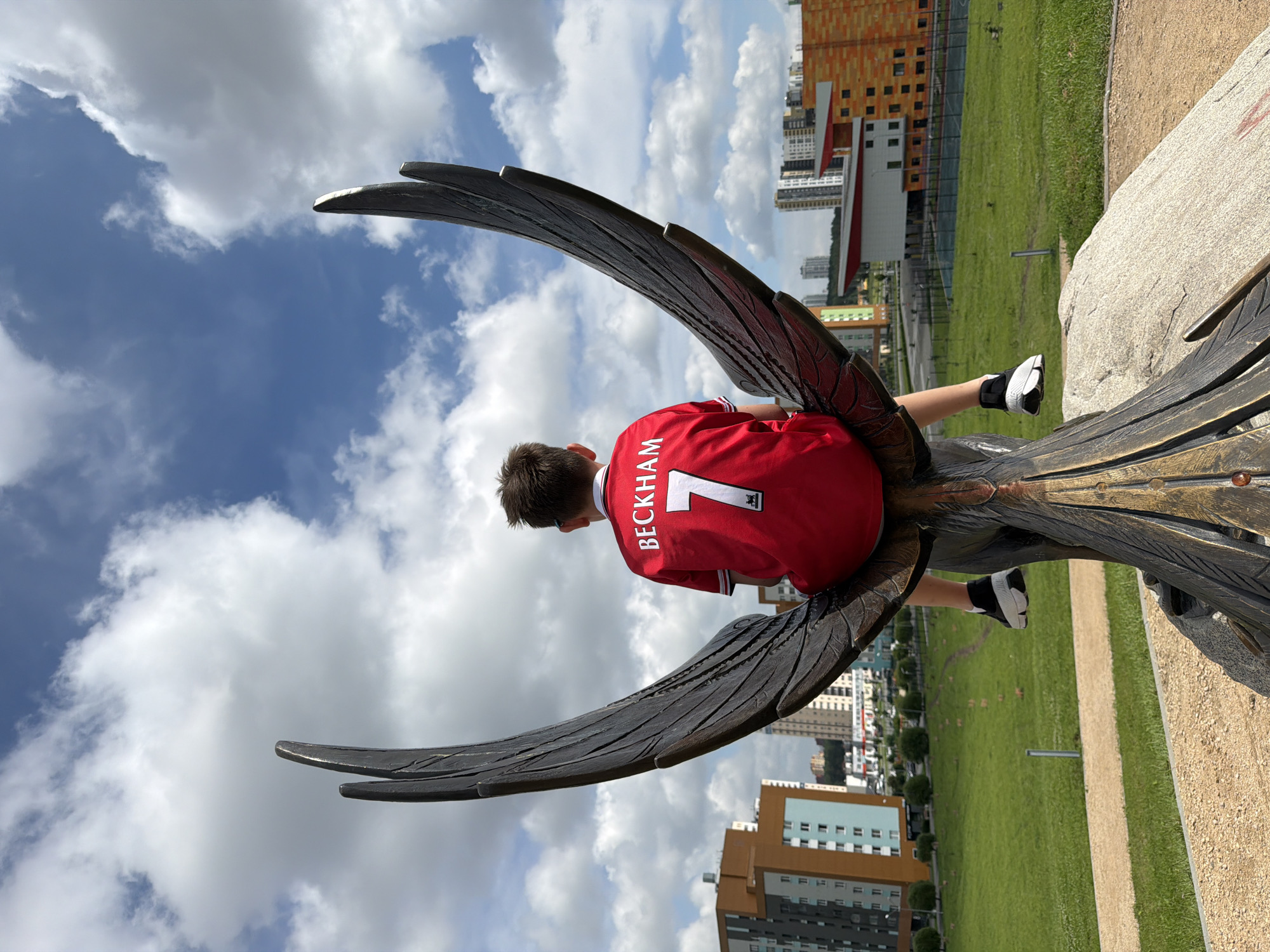 A young boy, seen from behind, sits on a large, dark metal sculpture resembling a bird's wings. He is wearing a red football jersey with "BECKHAM" and the number "7" on the back, along with black and white sneakers. The sculpture is situated on a light-colored rocky base. In the background, a green grassy field stretches out, with a pathway cutting through it. Beyond the field, there are modern buildings with colorful facades, including apartments and what appears to be a school or community center. The sky above is blue with fluffy white clouds. This scene is located in Yekaterinburg, Russia, specifically within the area of the Yekaterinburg City Administration's Children's Art Center.