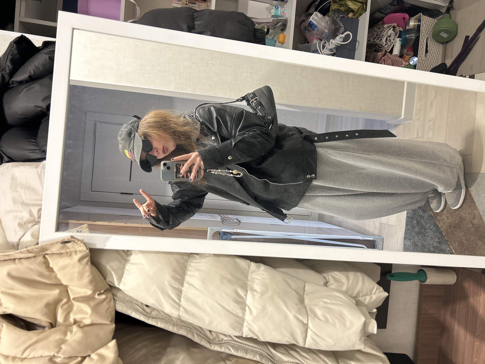A person is reflected in a rectangular white-framed mirror, holding a smartphone. The person is wearing a cap, sunglasses, a black leather jacket over a gray sweater, and gray sweatpants. A black purse is slung over the shoulder. The background includes clothing such as a black puffer jacket and a tan-colored comforter. A shelf filled with various items is visible above the mirror. A lint roller is located on the floor to the right of the mirror. The location is not identifiable as Krasnoyarsk, Russia based on the provided image.