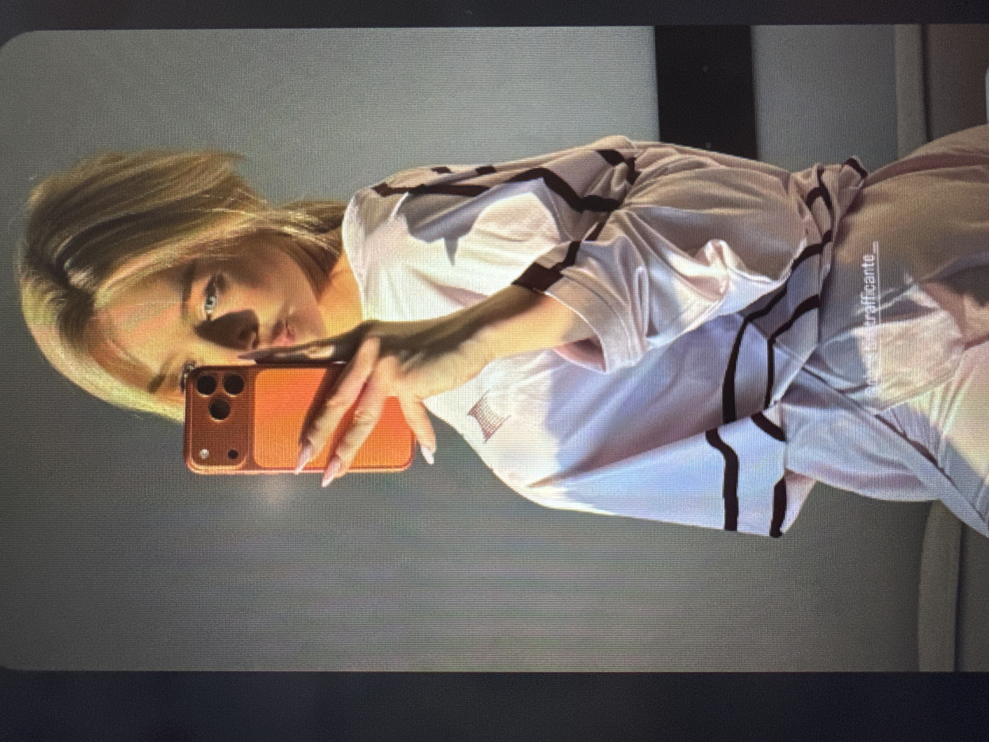 A young woman with blonde hair and blue eyes is captured in a selfie, likely taken in a mirror or reflective surface. She looks directly at the camera with a neutral expression, her hair styled to fall across her face. Her long, pointed nails are painted a light pink. She is wearing a light-colored, possibly pink or cream, long-sleeved top or robe featuring dark, geometric stripes. She holds an orange-colored smartphone, likely an iPhone, in her right hand, positioned to take the picture.
The background is a plain, light gray wall, suggesting an indoor setting, possibly a room. A darker shadow or object is visible on the left side of the background. The lighting is soft and directional, creating subtle highlights and shadows on her face and clothing.
Visible on her right sleeve is the text "@trafficante_". The image displays a slight graininess, possibly indicating it is a photograph of a screen. The overall scene conveys a casual and relaxed atmosphere.