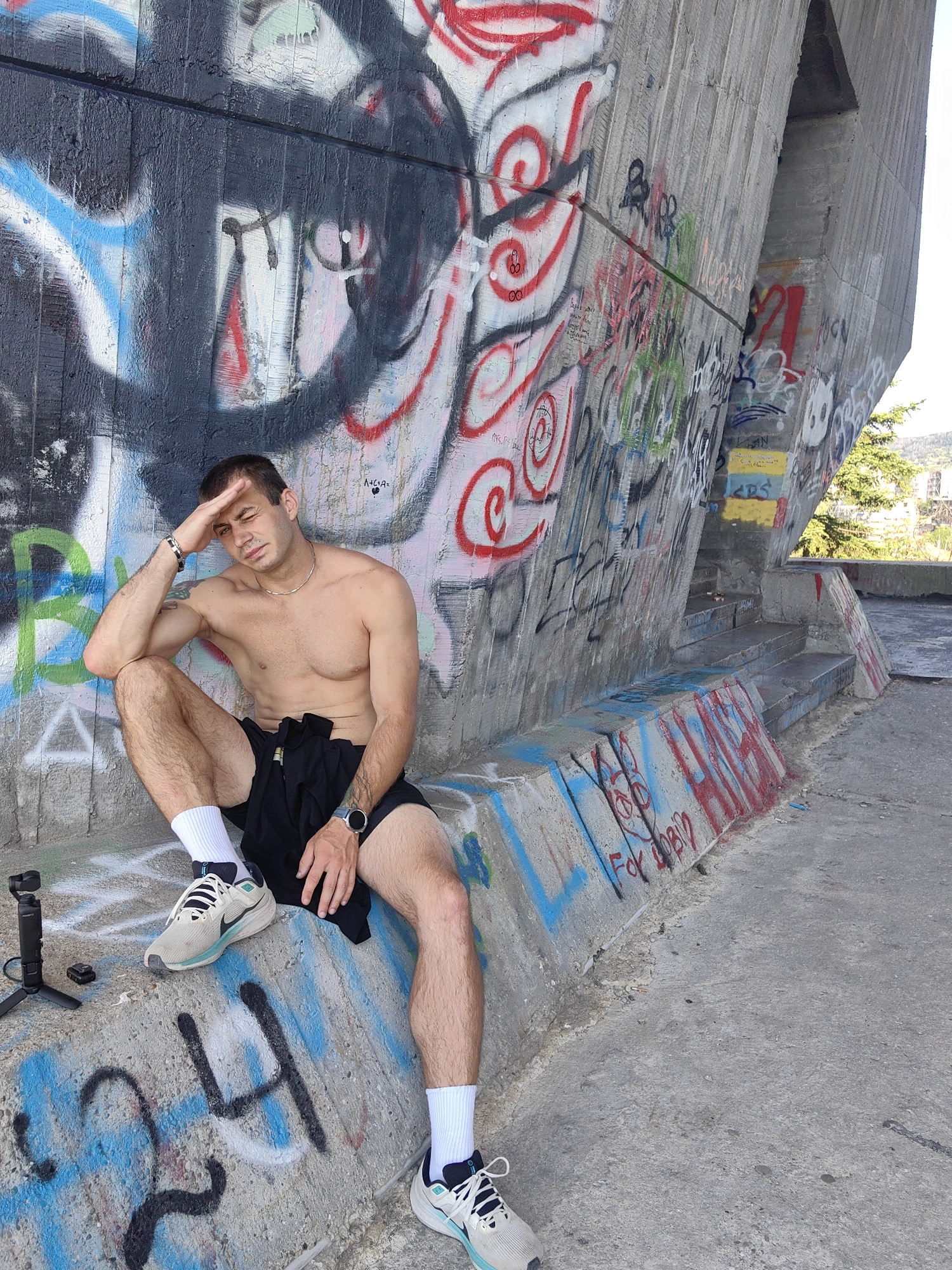 A shirtless young man with short brown hair, wearing black shorts, white socks, and grey and white athletic shoes, is sitting on a concrete ledge against a graffiti-covered concrete wall. He is leaning on his left arm with his right hand resting on his forehead. A black gimbal stabilizer and a small black device are on the ledge to his left. The graffiti on the wall is colorful, with various tags and designs. The ground is also concrete. In the background, a green tree and some buildings are visible.
