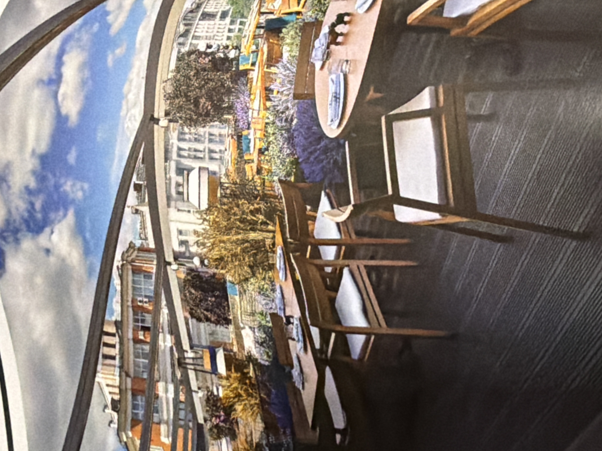 The image depicts an outdoor restaurant in a city. The restaurant appears to be situated on a rooftop or elevated platform, offering a view of the surrounding cityscape. The furniture consists of wooden tables set with plates and cutlery, and chairs with white cushions. The setting includes decorative plants, such as lavender and other greenery, adding to the ambiance. A metal framework arches over the restaurant area, giving it a tent-like appearance, with a blue sky visible above. The city buildings are visible in the background, suggesting a metropolitan environment. No people are visible. It is believed that the image is of an outdoor restaurant in Moscow, Russia.