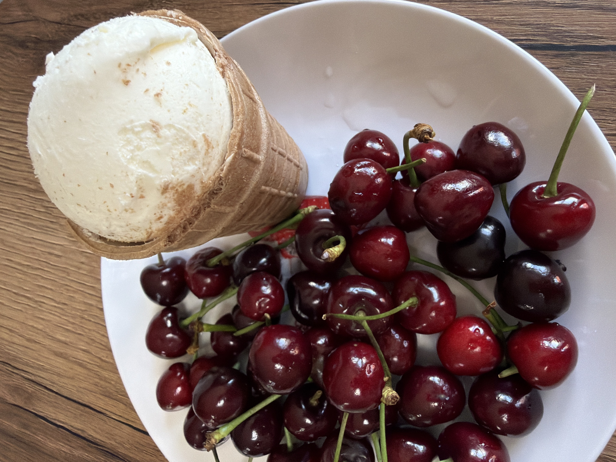 The image shows a white plate with a pile of fresh cherries and a scoop of vanilla ice cream in a waffle cone. The plate rests on a wooden table. The cherries are dark red and appear to be fresh and ripe, with some still attached to their green stems. The ice cream is a pale yellow-white color and has a slightly grainy texture, suggesting it might contain vanilla bean specks. The waffle cone is light brown and has a classic criss-cross pattern. The lighting is natural and soft, casting gentle shadows on the table. The background is out of focus, but the texture of the wooden table is visible. There is no indication of a specific location in the city of Chaikovsky, Russia; the scene is a generic still life arrangement.